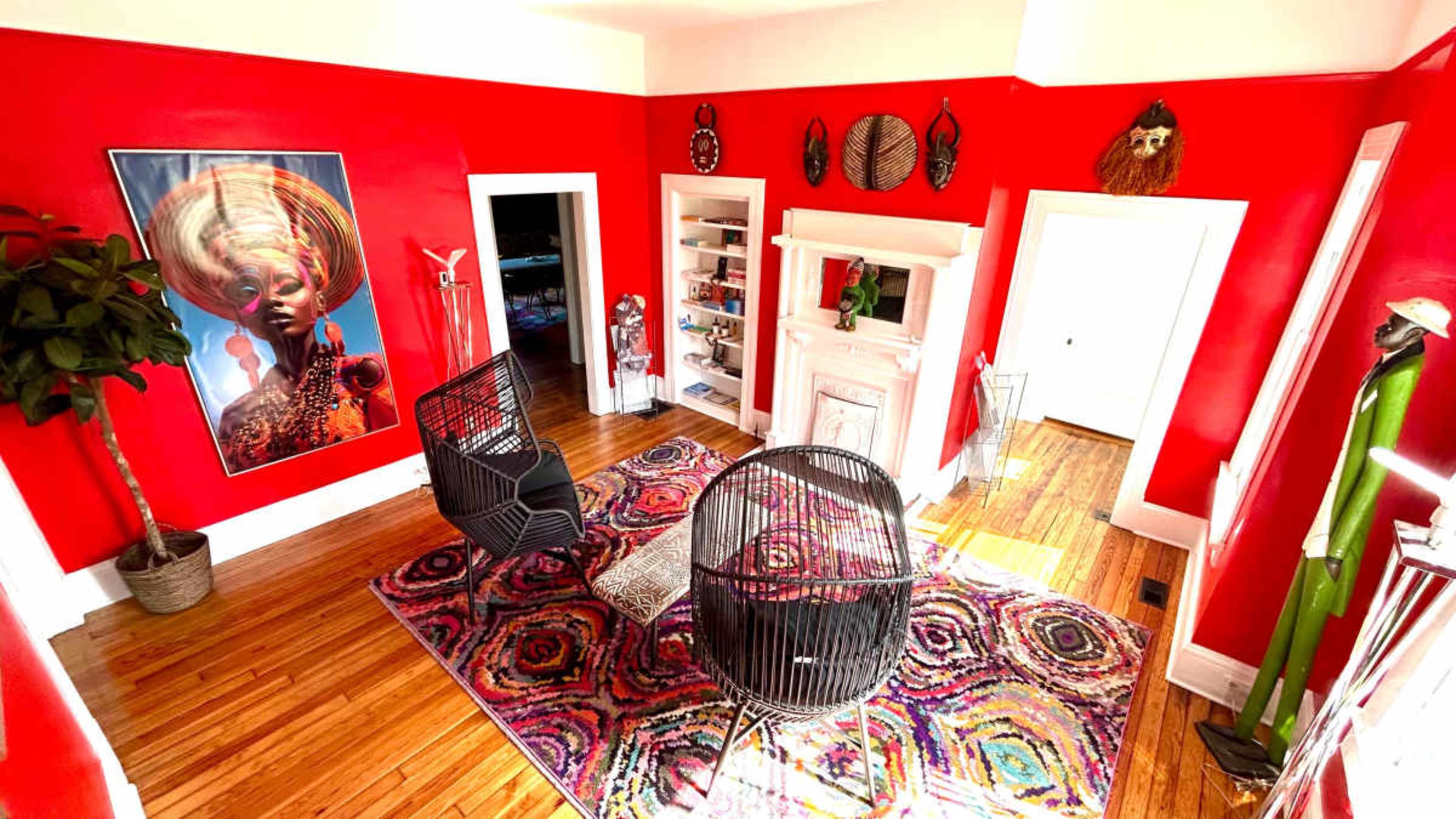The room features bright red walls, a large colorful rug, two black woven chairs, and various decorative items on the shelves and walls.
