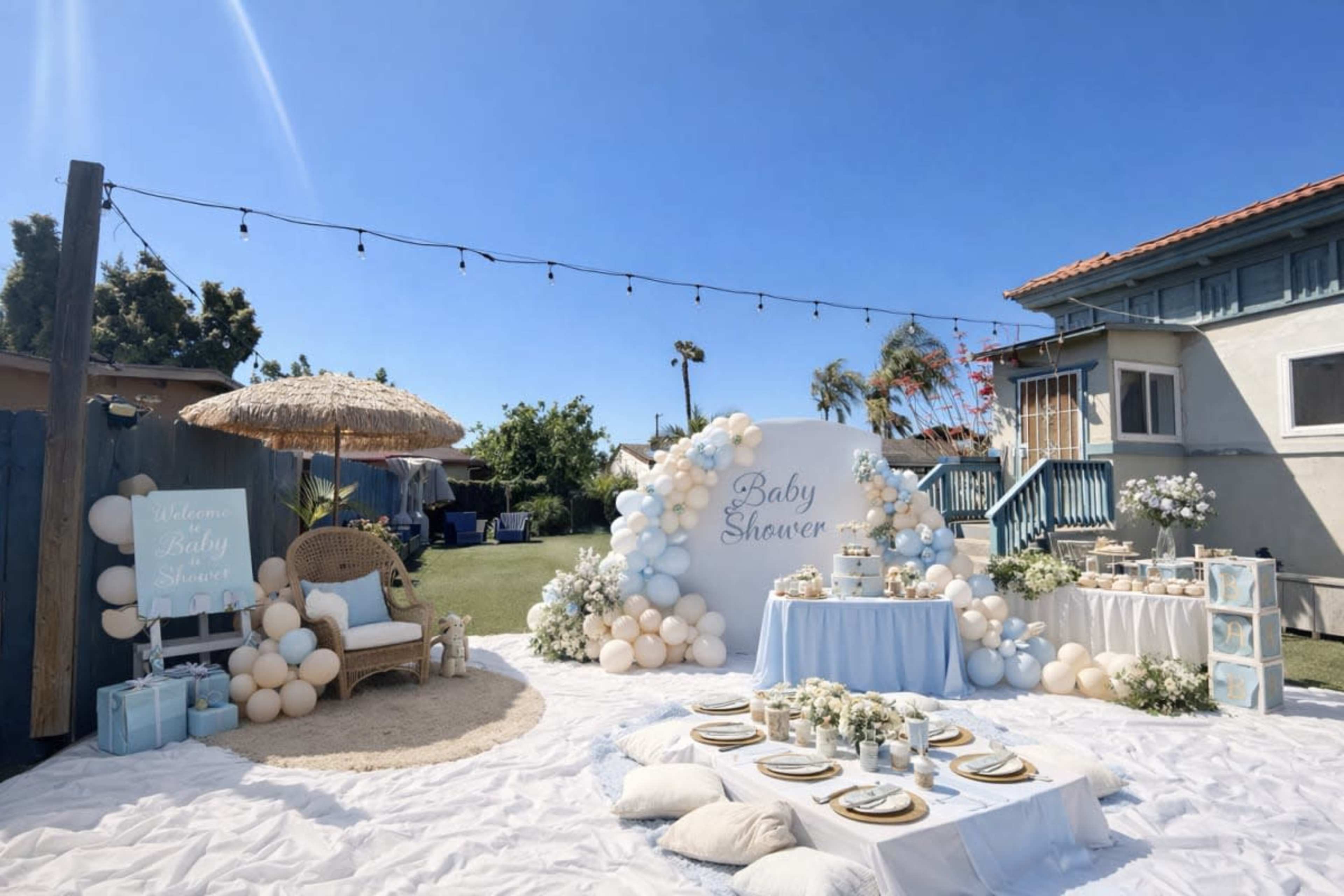 The image shows a decorative outdoor space set up for a baby shower, featuring a blue and white color scheme with a table, balloon arrangements, and seating under a thatched umbrella.