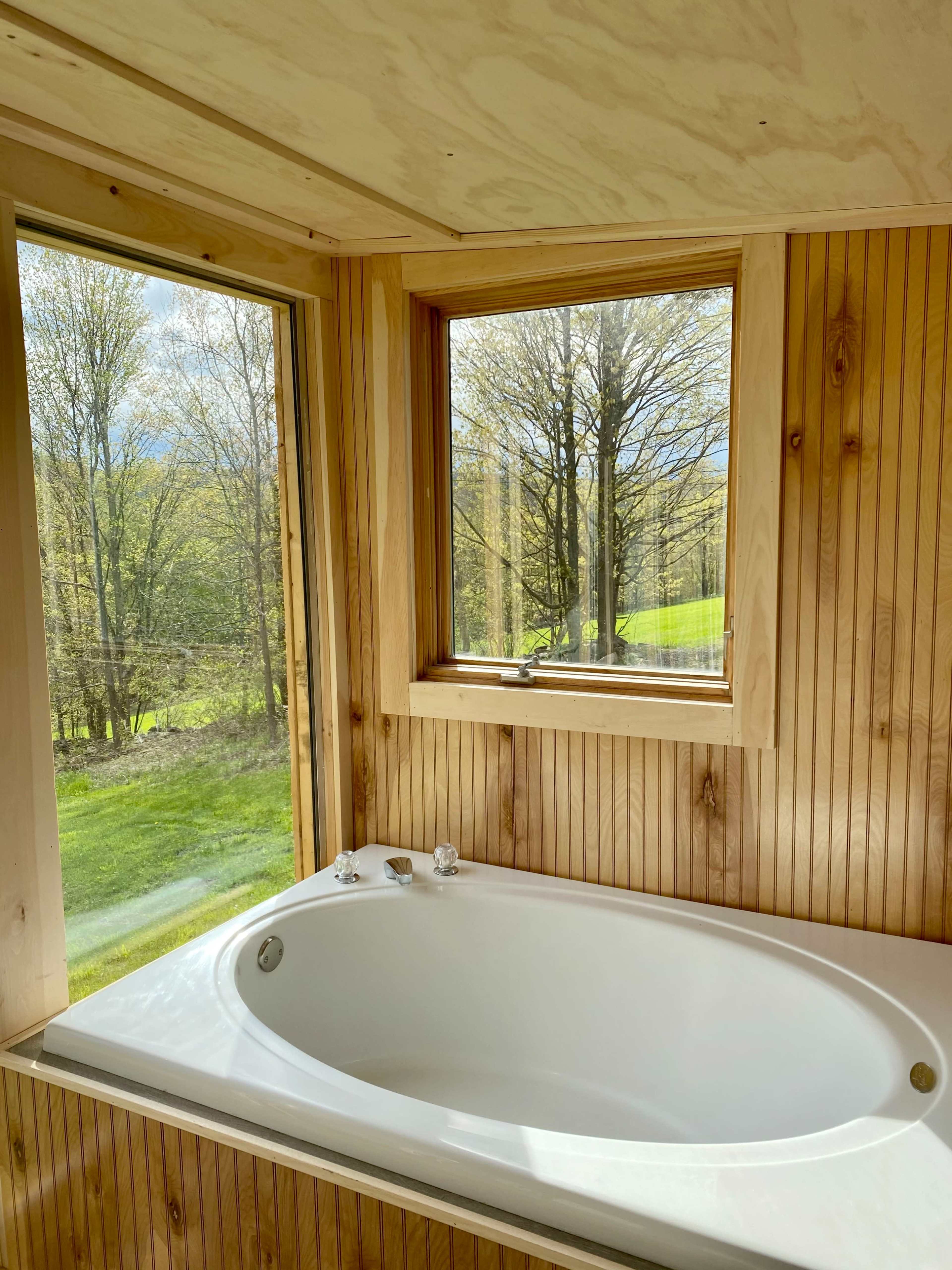 A white bathtub is situated in a wooden bathroom with large windows showcasing a view of trees and grassy landscape outside.