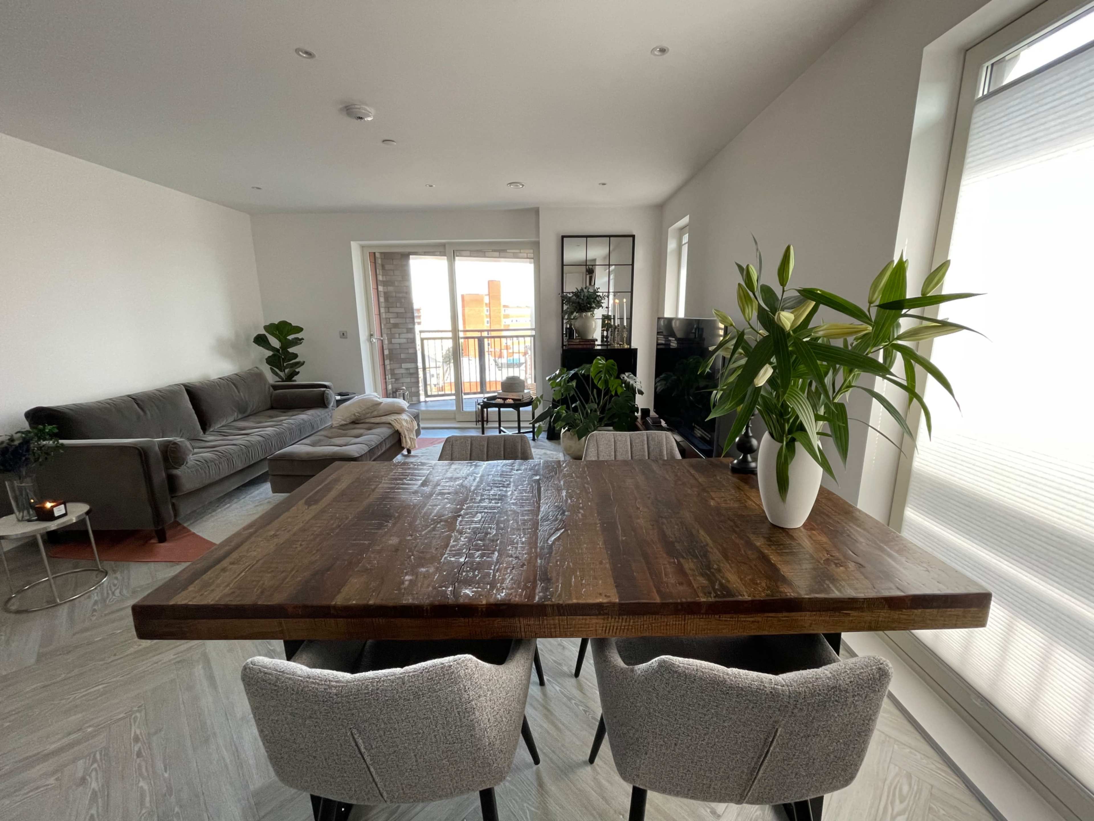 A wooden dining table with gray chairs is positioned in the foreground, while a living room featuring a gray sofa and large windows is visible in the background.