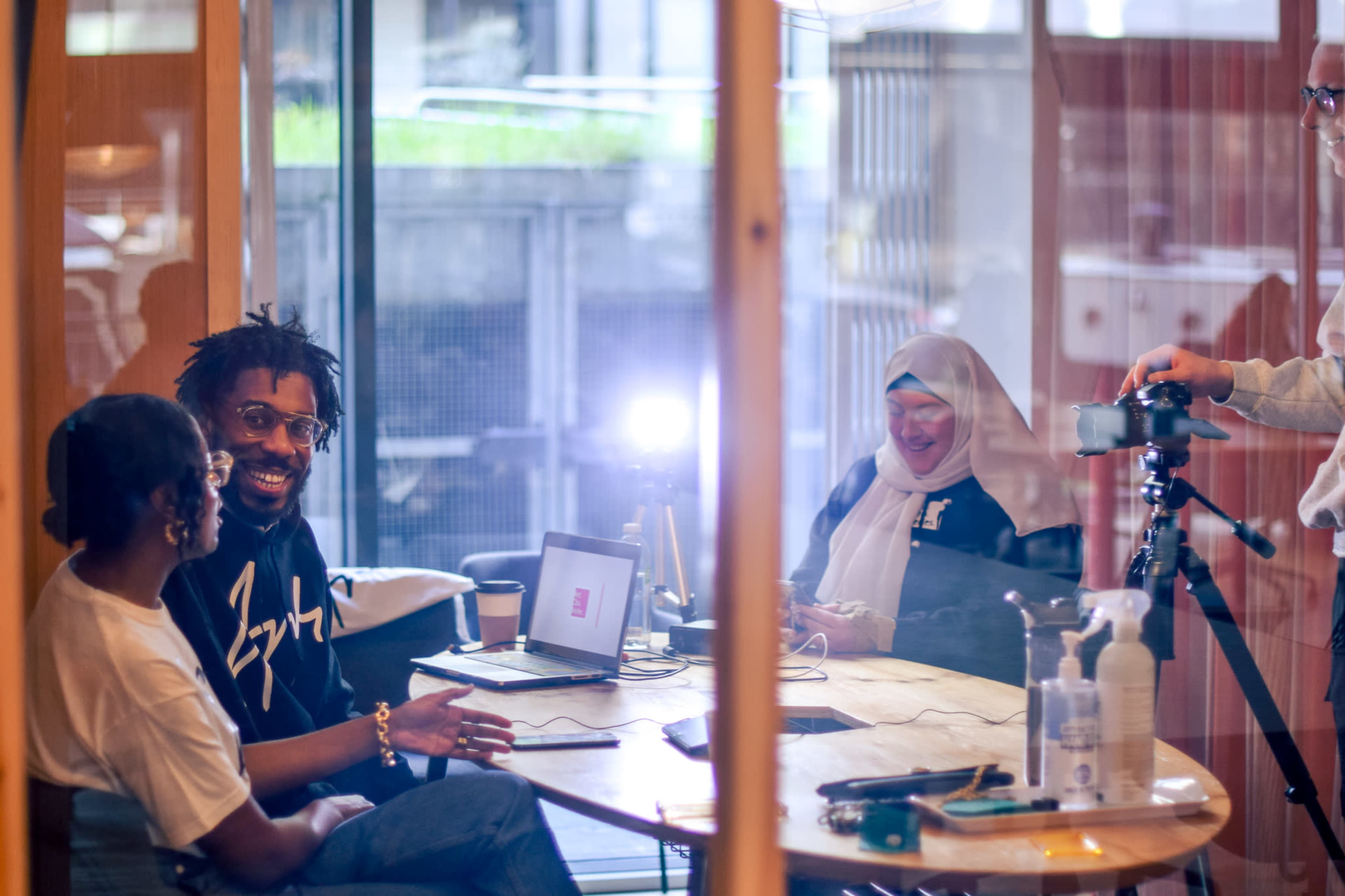 A group of three people is engaged in a conversation around a table while a fourth person sets up a camera in a brightly lit room.