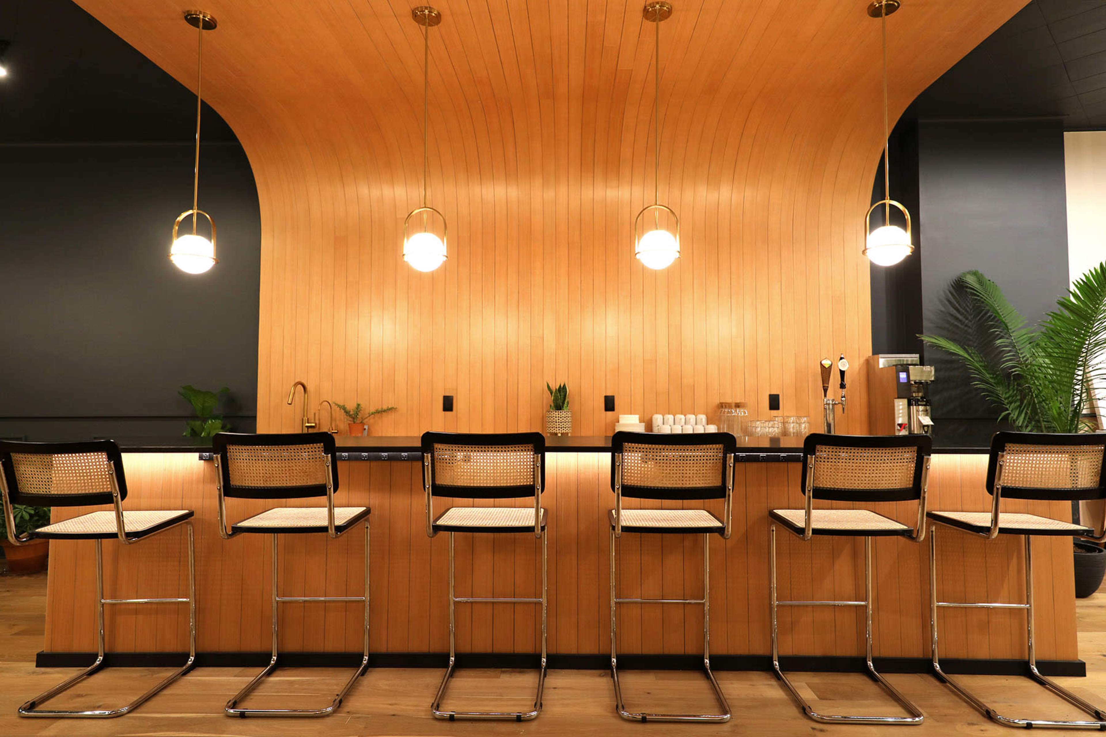 A wooden bar with a curved design features several high stools lined up in front of it and pendant lighting overhead.