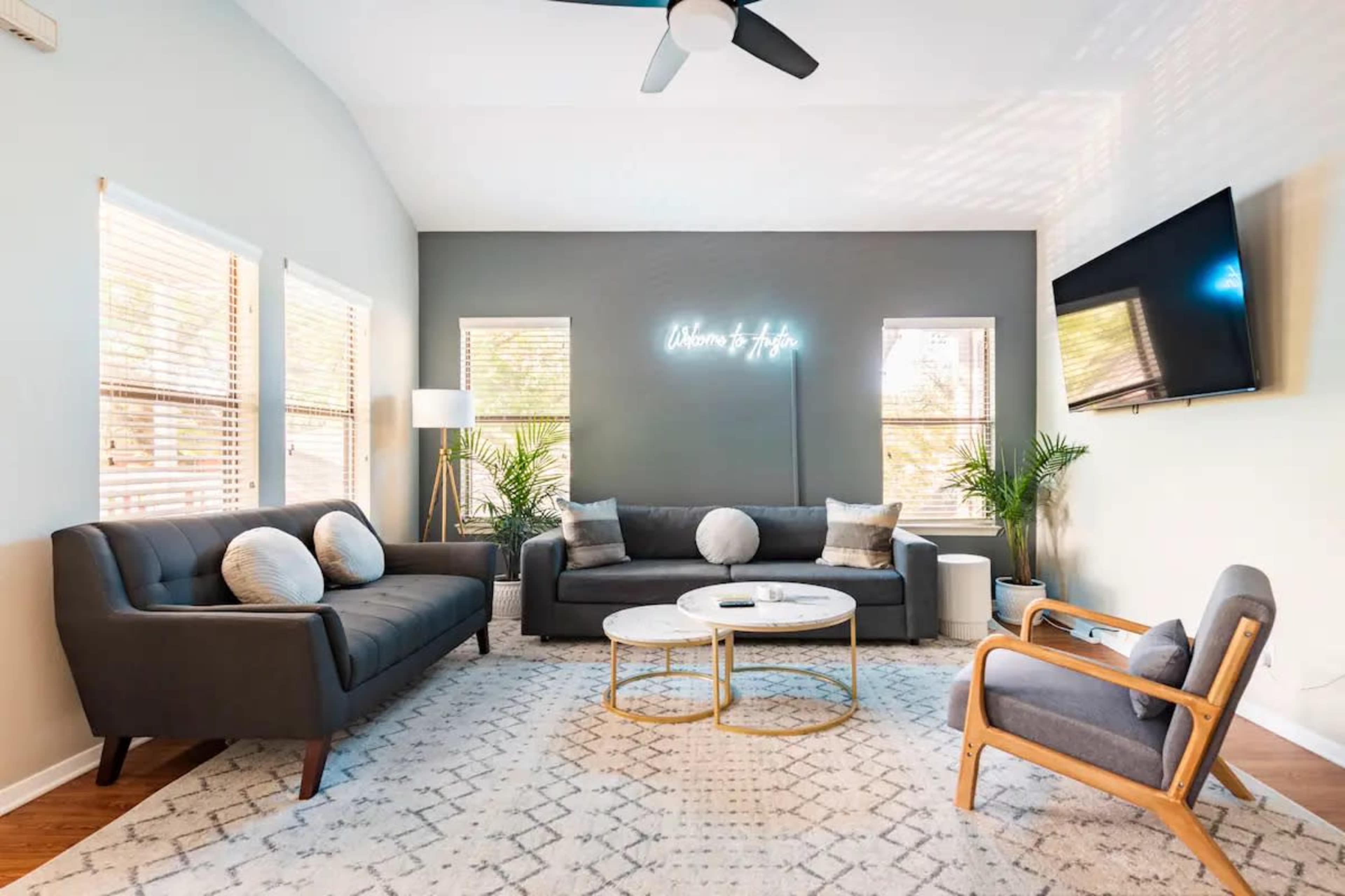 The living room features two dark sofas, a round coffee table, and a television mounted on the wall, with large windows allowing natural light to enter.
