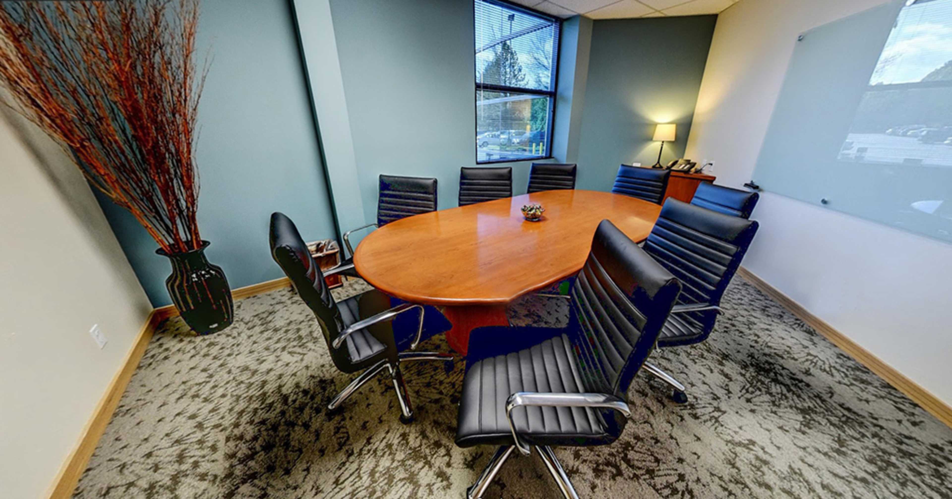 A conference room features an oval wooden table surrounded by black leather chairs, with a vase and a small plant on the table.