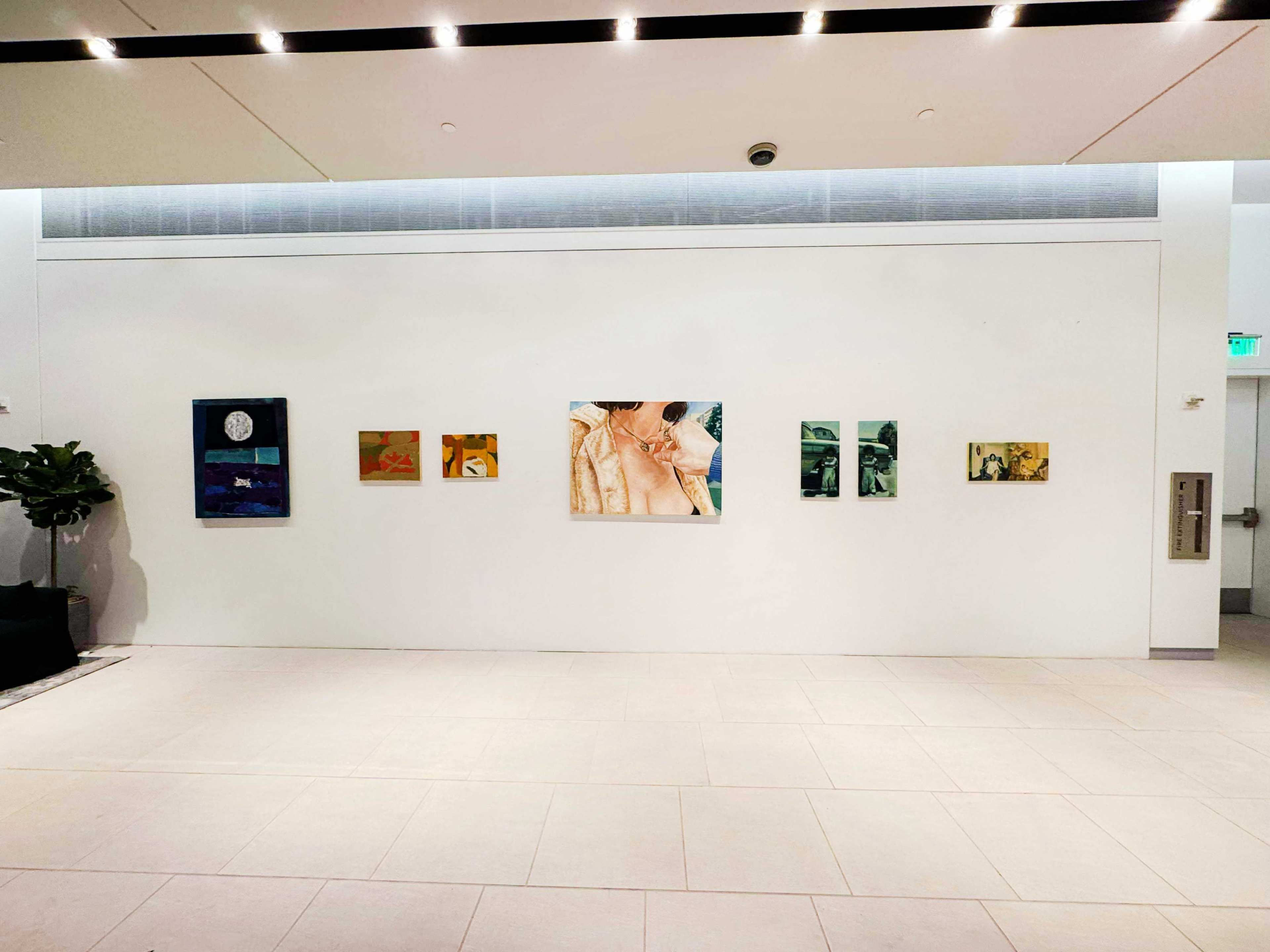 The image shows a white wall in a gallery or modern space with six pieces of art displayed in a row, alongside a potted plant in the corner.