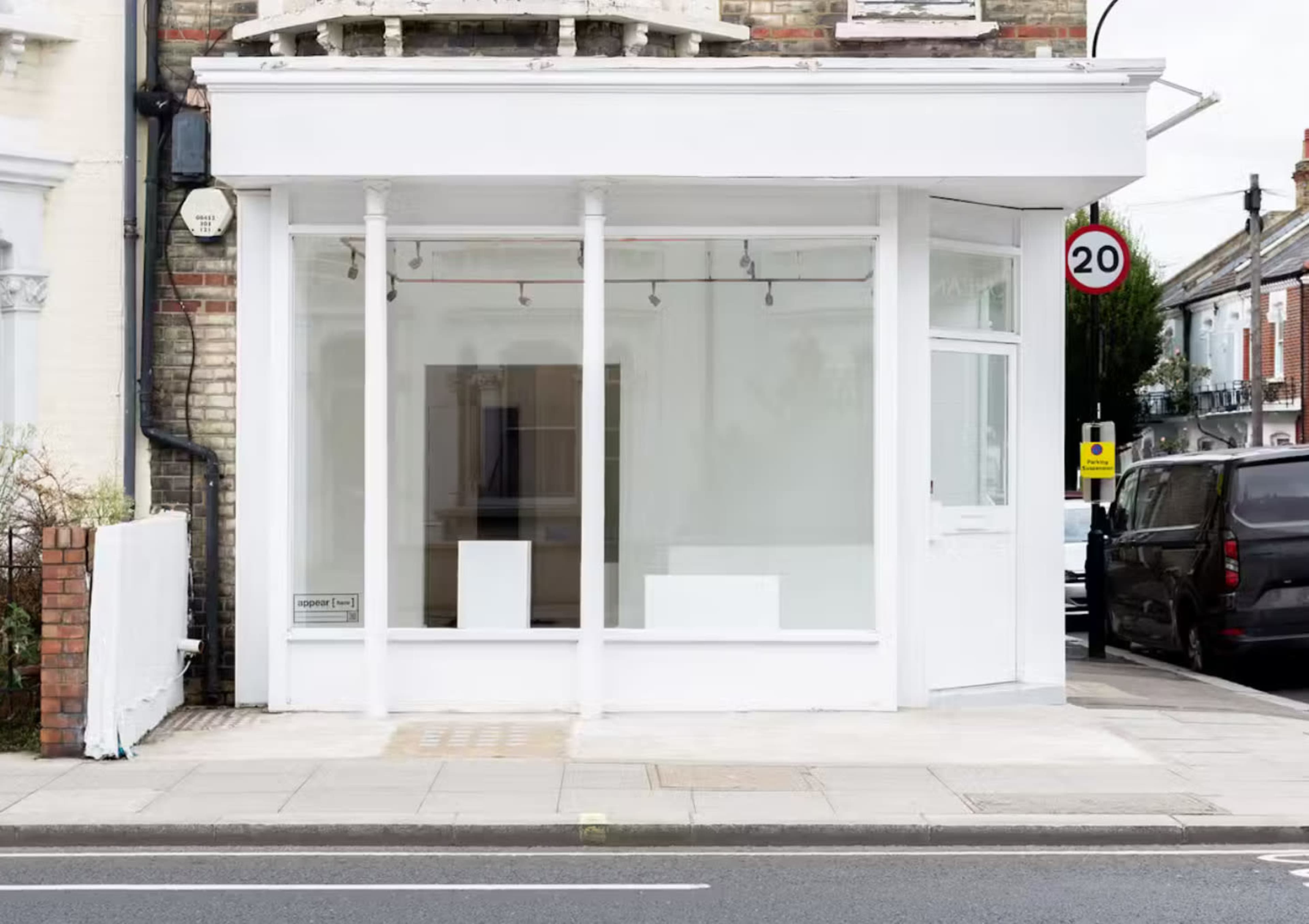The image shows a vacant retail space with large glass windows and a white exterior, located on a street with parked cars and a traffic sign nearby.