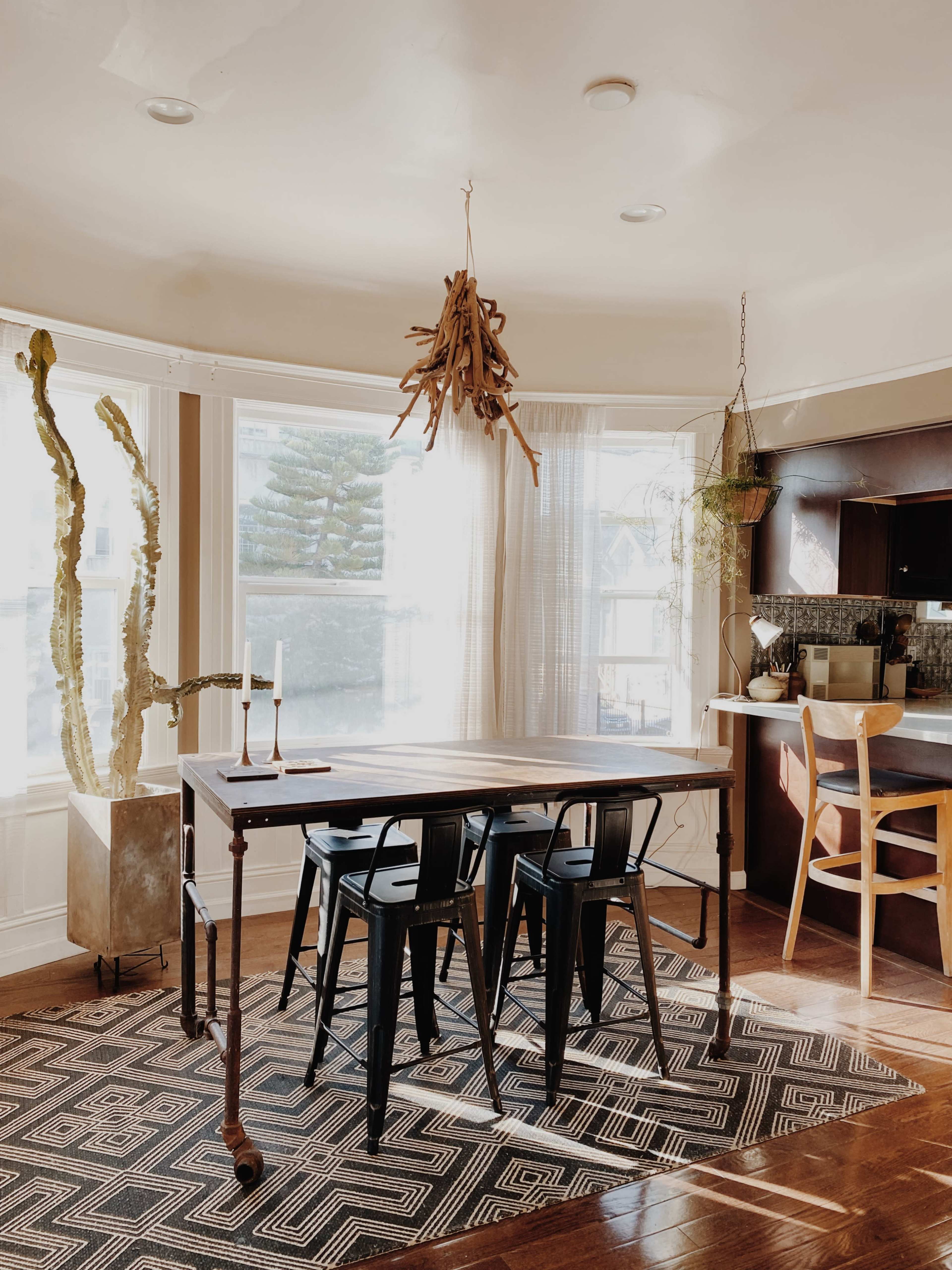A dining area with a wooden table, black metal chairs, a cactus in a planter, and a kitchen visible in the background.