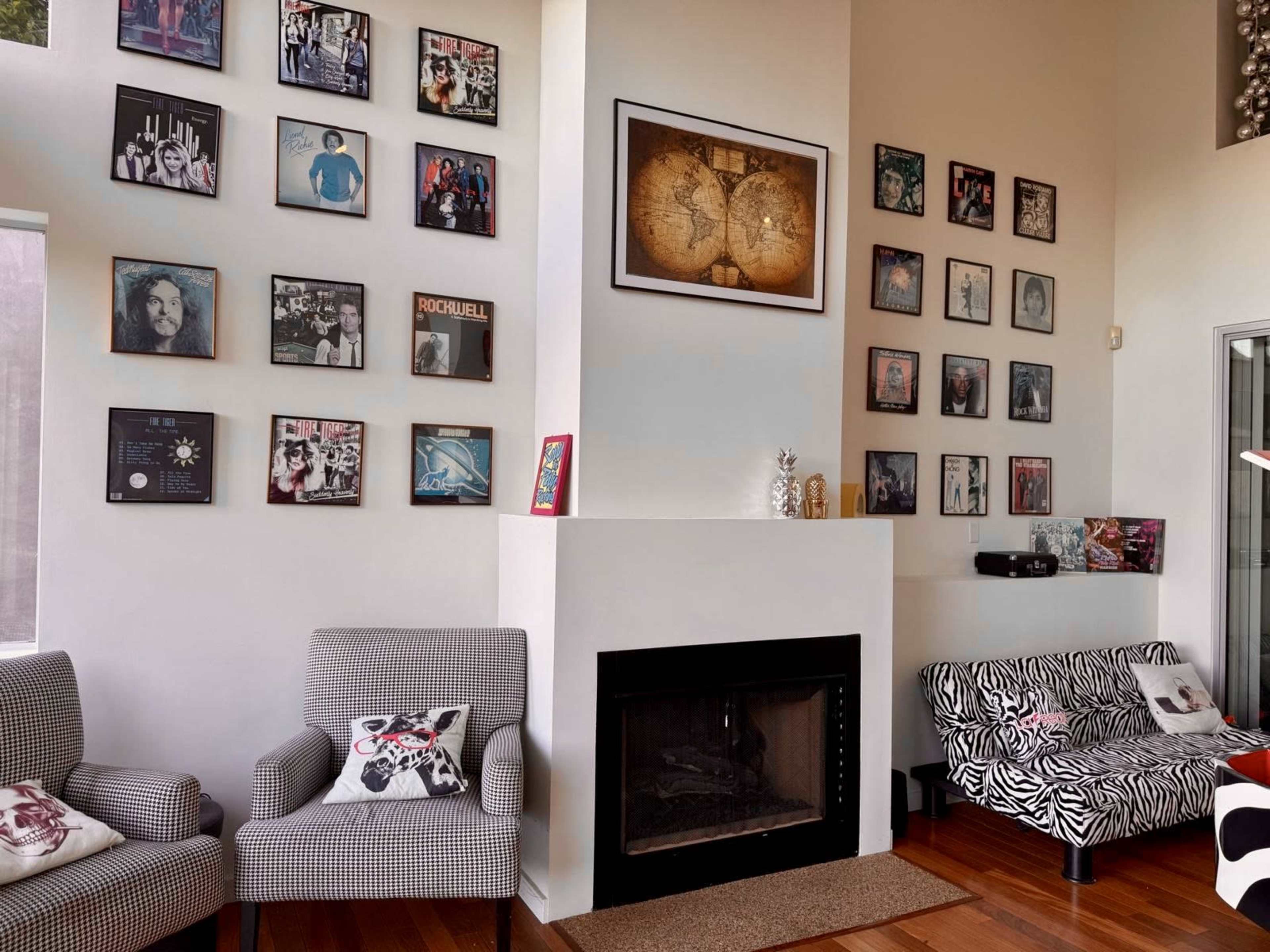 The image shows a living room featuring a fireplace, two patterned chairs, a striped sofa, and a wall adorned with various framed pictures and vinyl records.
