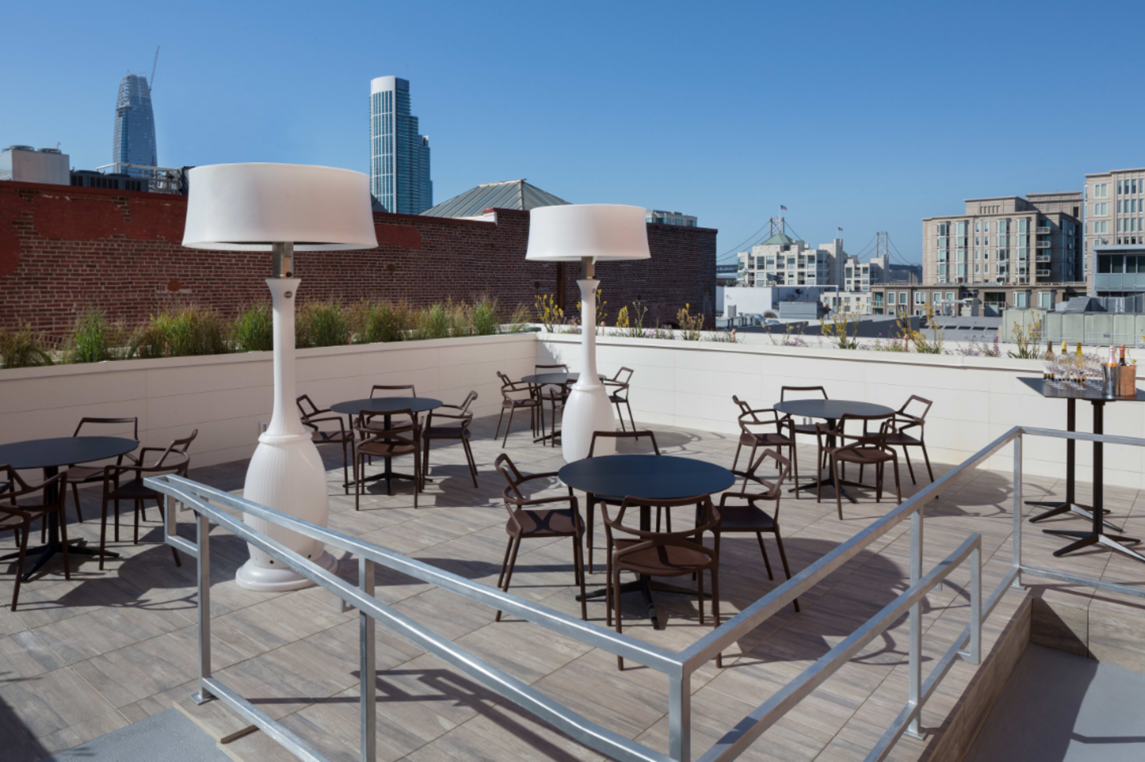 A rooftop terrace with several round tables and chairs, surrounded by a railing, and two large white lamp structures, set against a clear blue sky and a city skyline in the background.