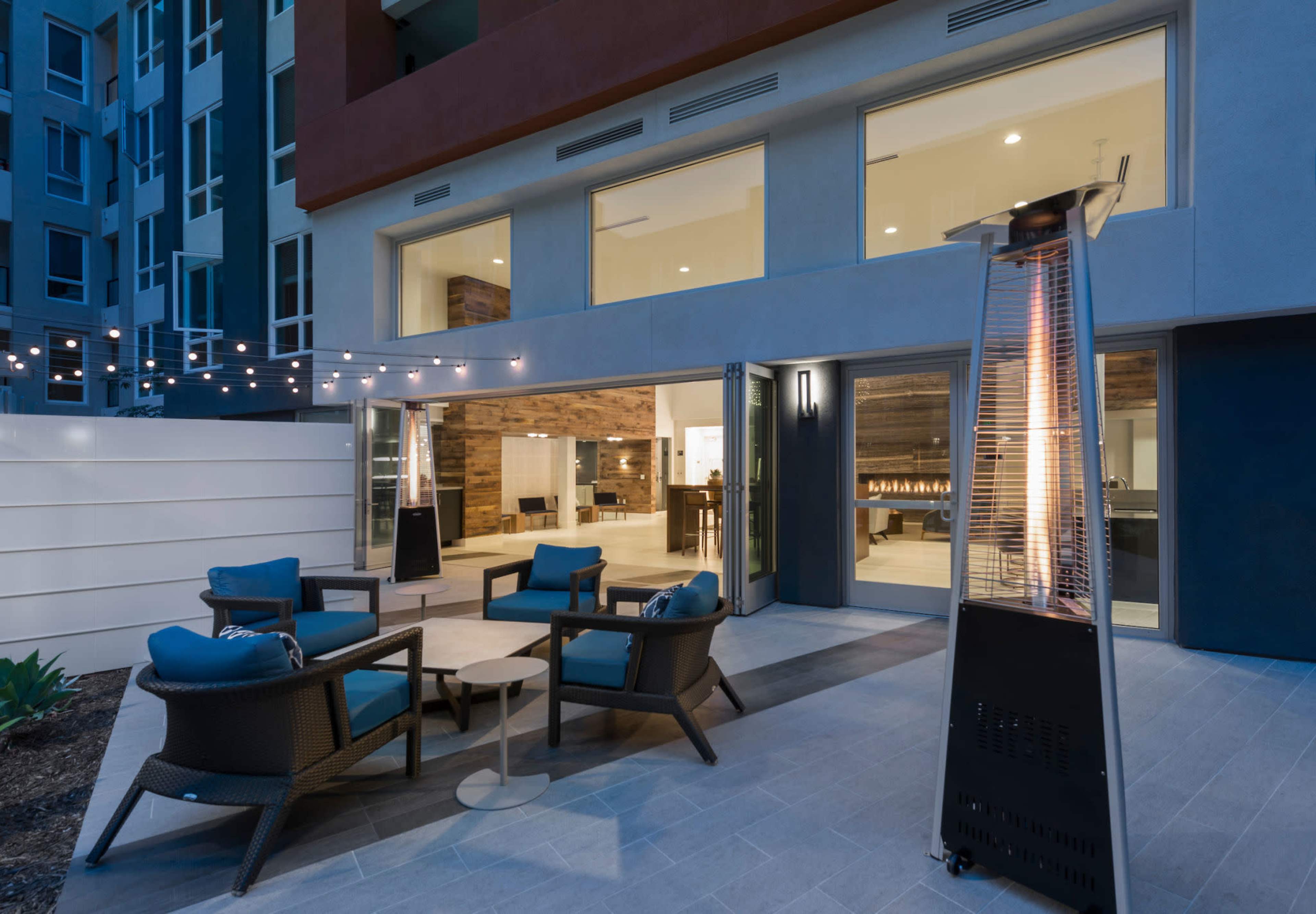 A modern outdoor patio area with seating, a table, and a gas patio heater, surrounded by buildings and string lights.