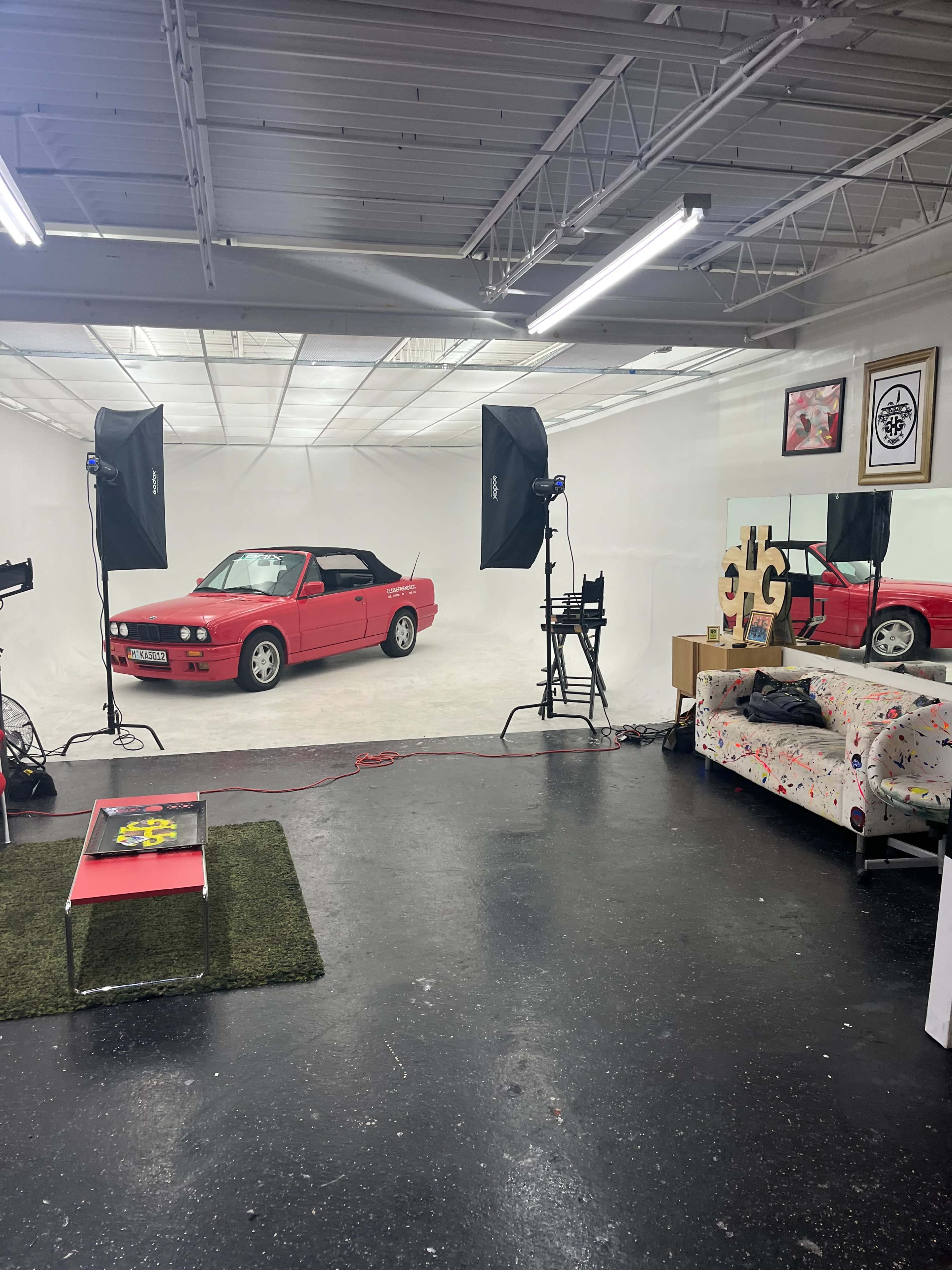 The image shows a bright photo studio with a red car positioned in the center, surrounded by photography equipment and a colorful couch in the foreground.
