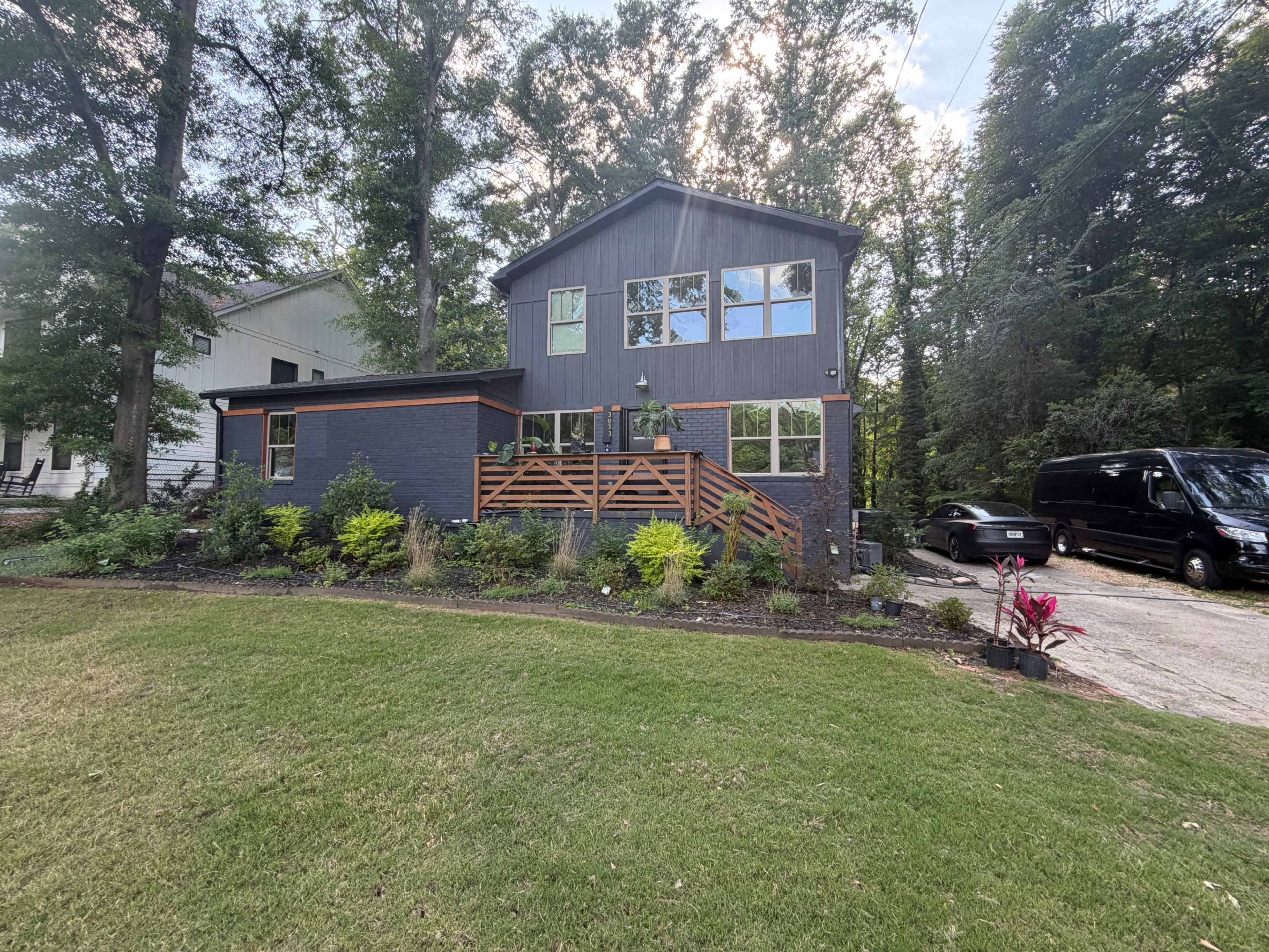 A modern two-story house with a dark exterior and large windows is surrounded by greenery and features a wooden deck in the front.