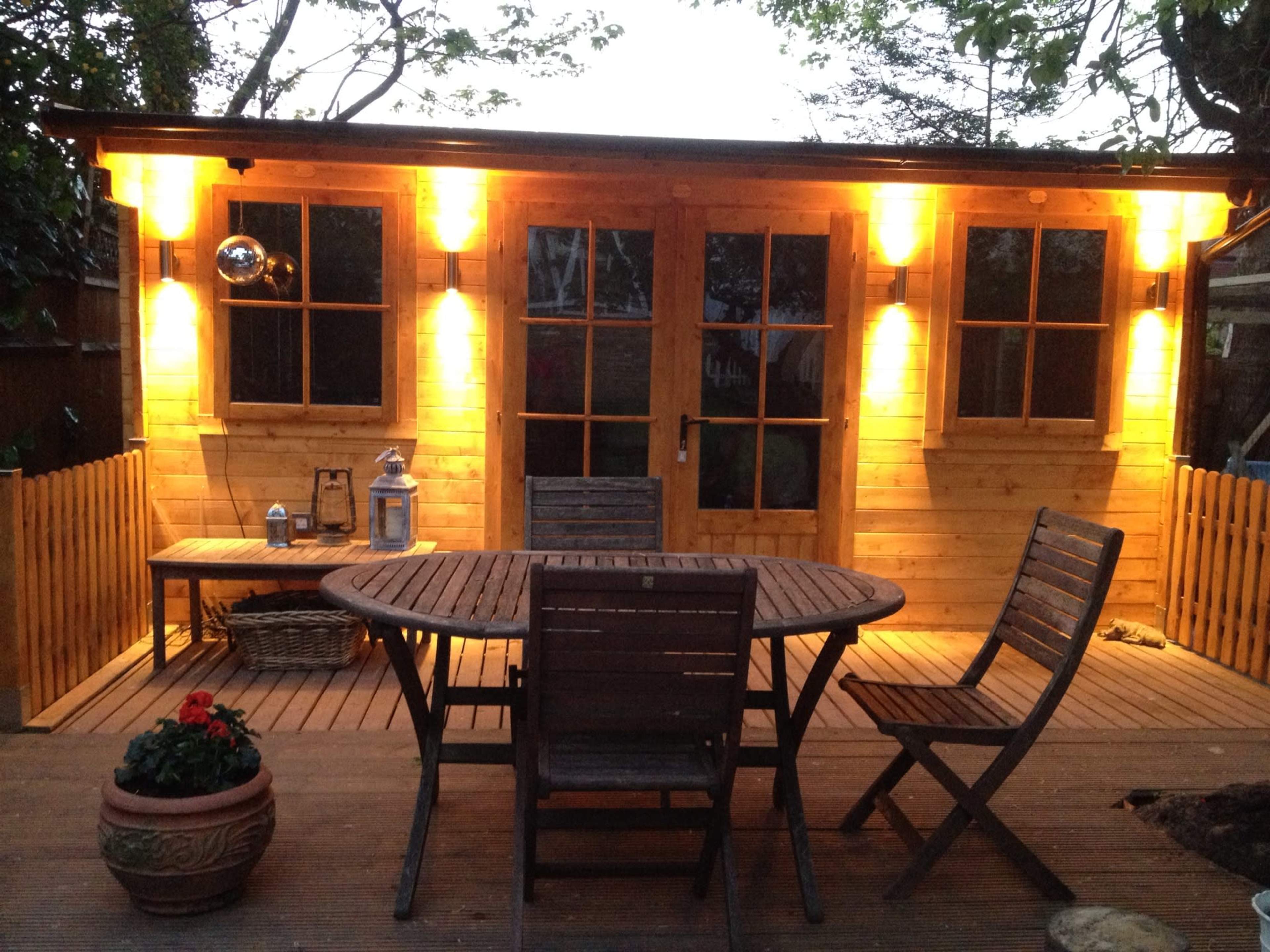 A wooden cabin with illuminated exterior lights is seen beside a circular wooden table and chairs on a deck, surrounded by greenery.