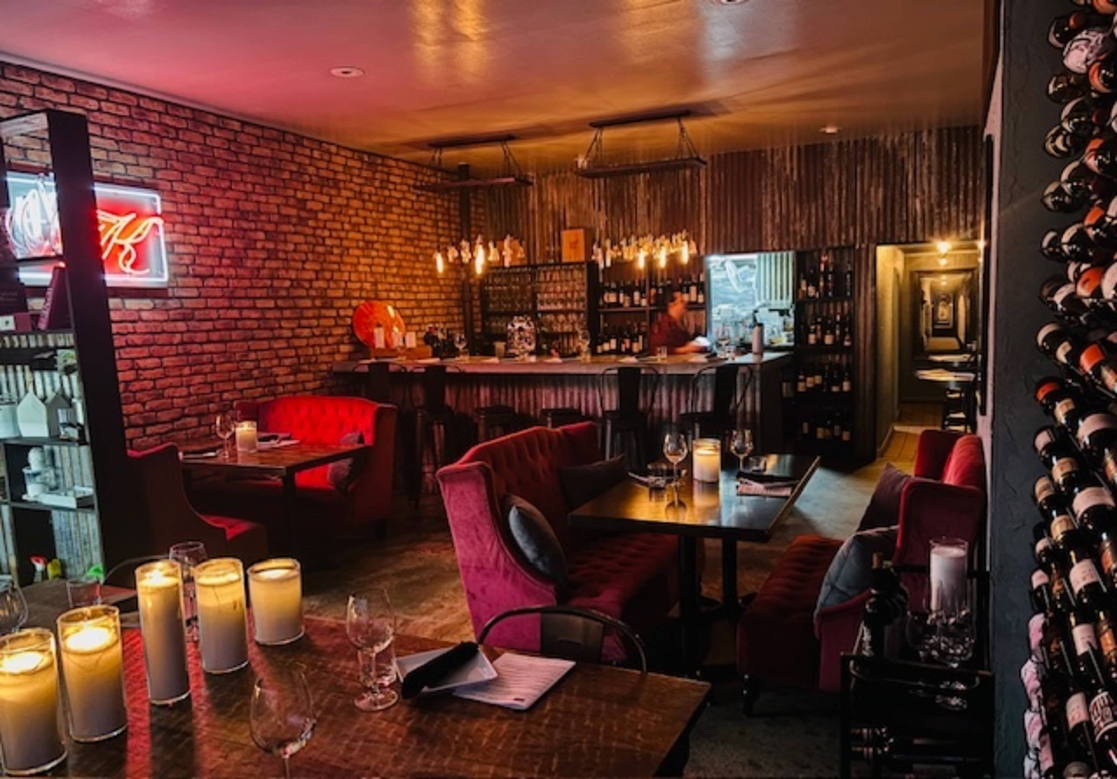 The image shows a cozy restaurant interior with brick walls, plush red seating, a bar area with bottles, and soft lighting from candles and hanging fixtures.