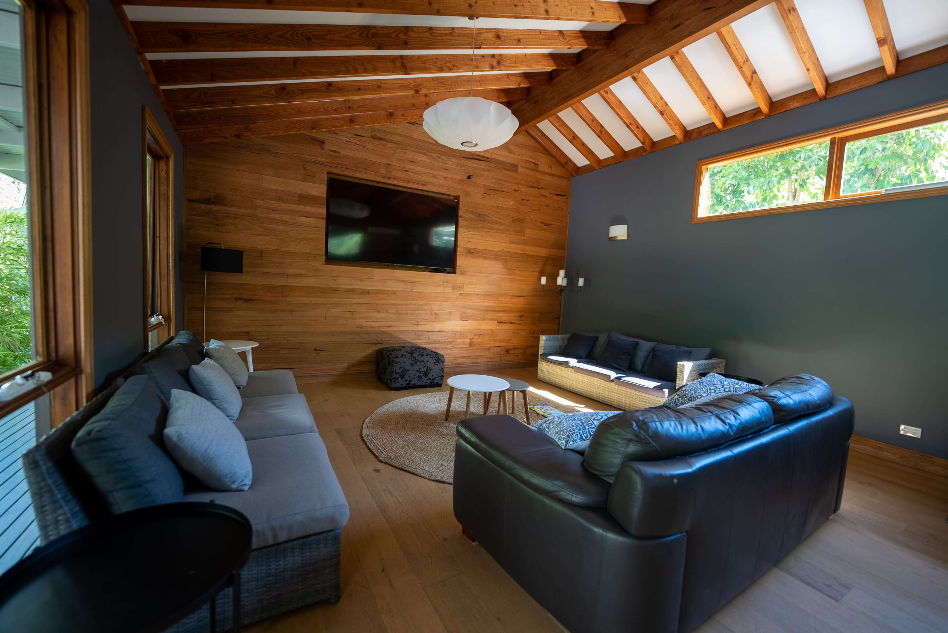 The image shows a living room with wooden walls, a high ceiling, and two seating areas, including a dark leather couch and a light-colored sofa.