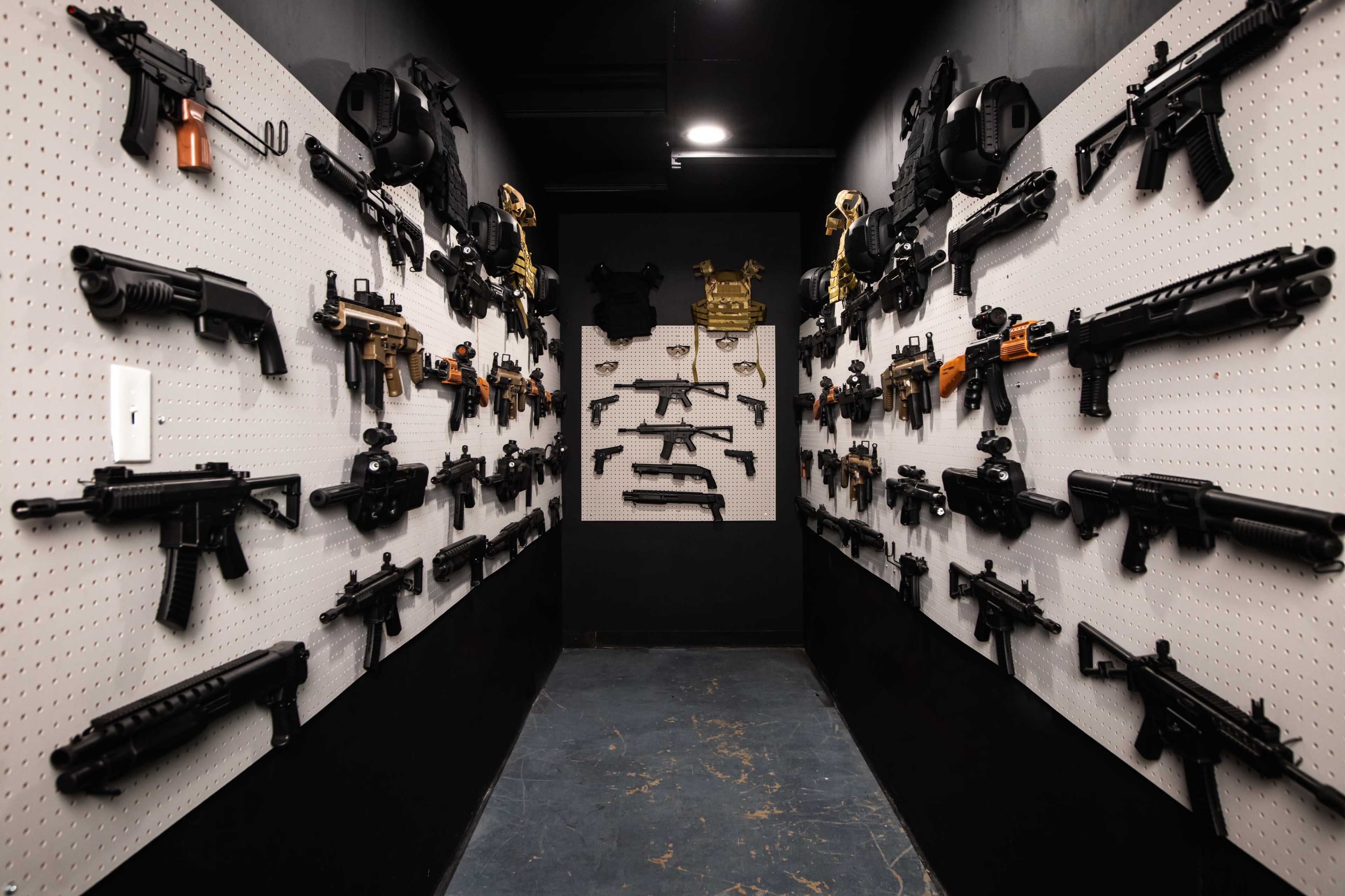 The image shows a narrow hallway lined with various firearms mounted on pegboards on both walls.