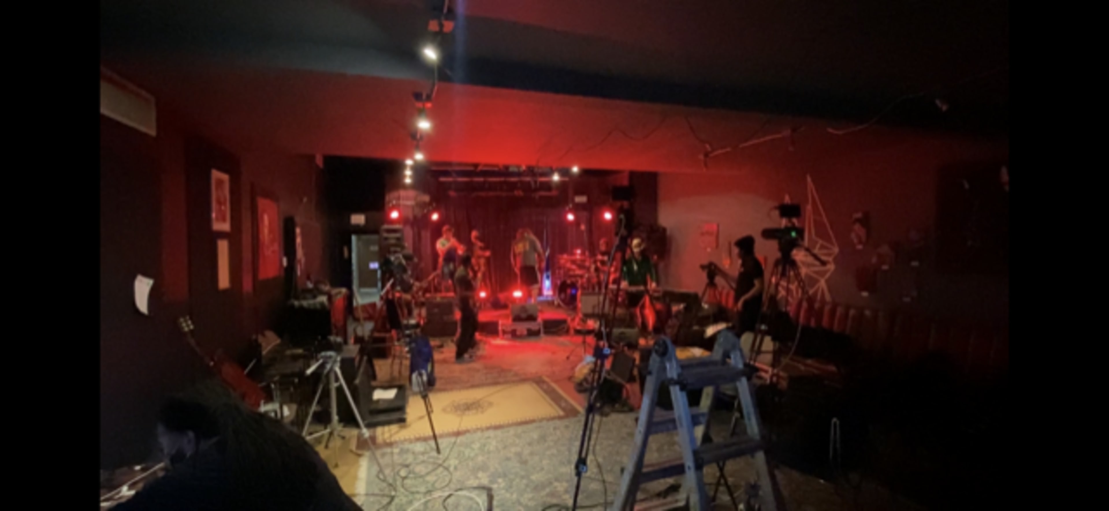 The image shows a dimly lit music venue set up for a performance, featuring red stage lights, musical instruments, and equipment arranged on the floor.