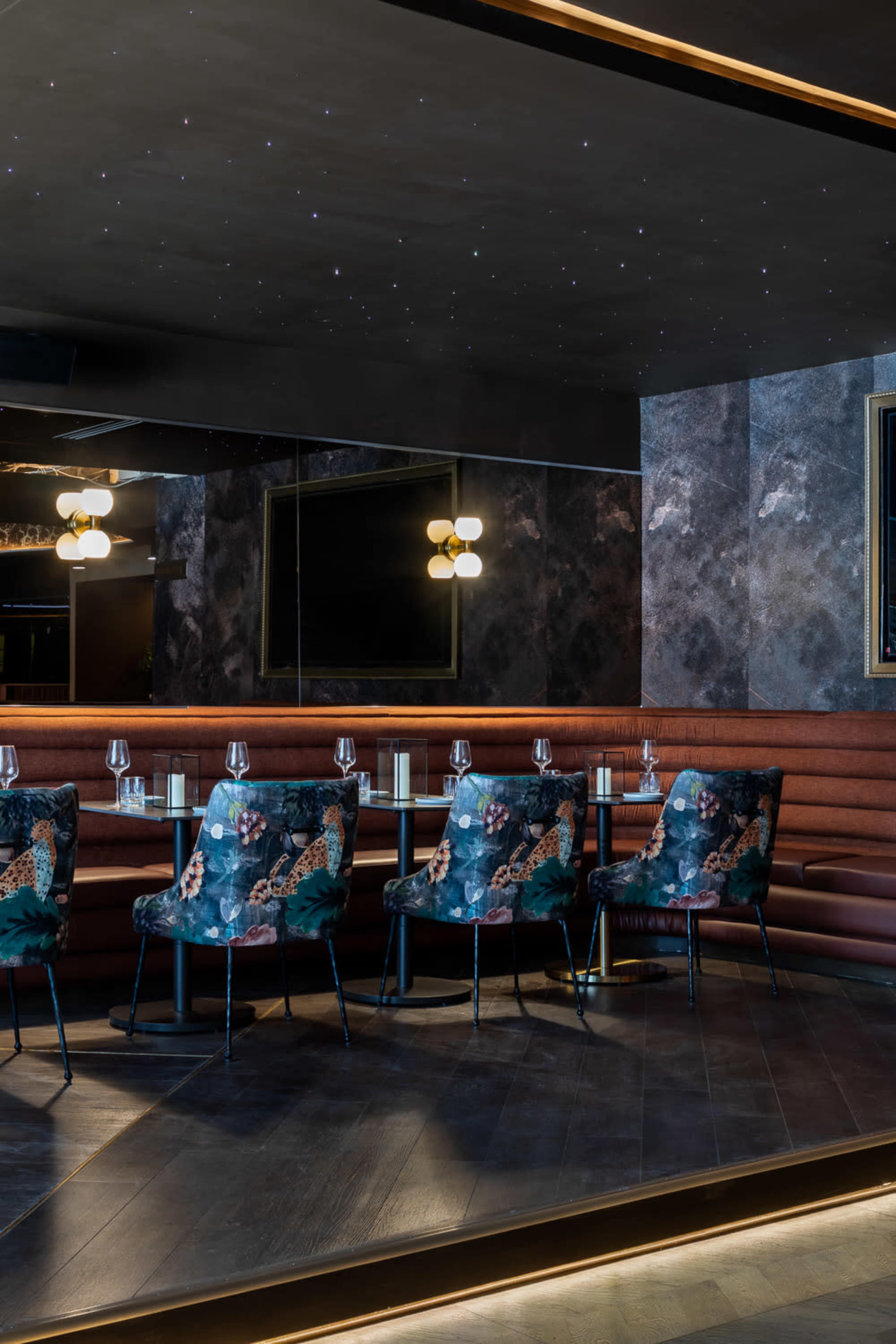The image shows a dimly lit restaurant interior featuring a mirrored wall, plush seating in deep colors, and floral patterned chairs arranged around a long table.