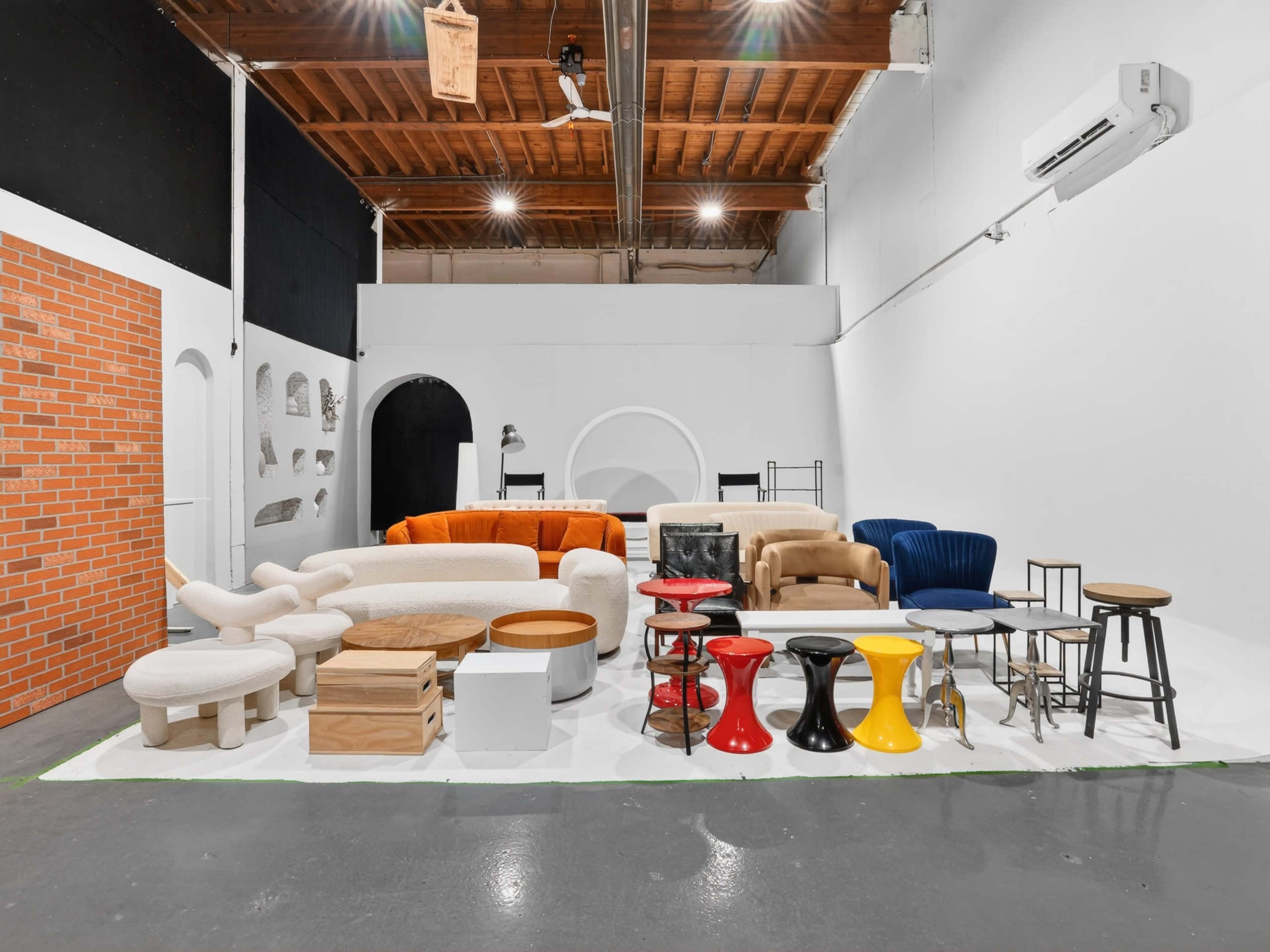 The image shows a variety of furniture pieces, including sofas, stools, and tables, arranged in a spacious indoor setting with a minimalist backdrop.