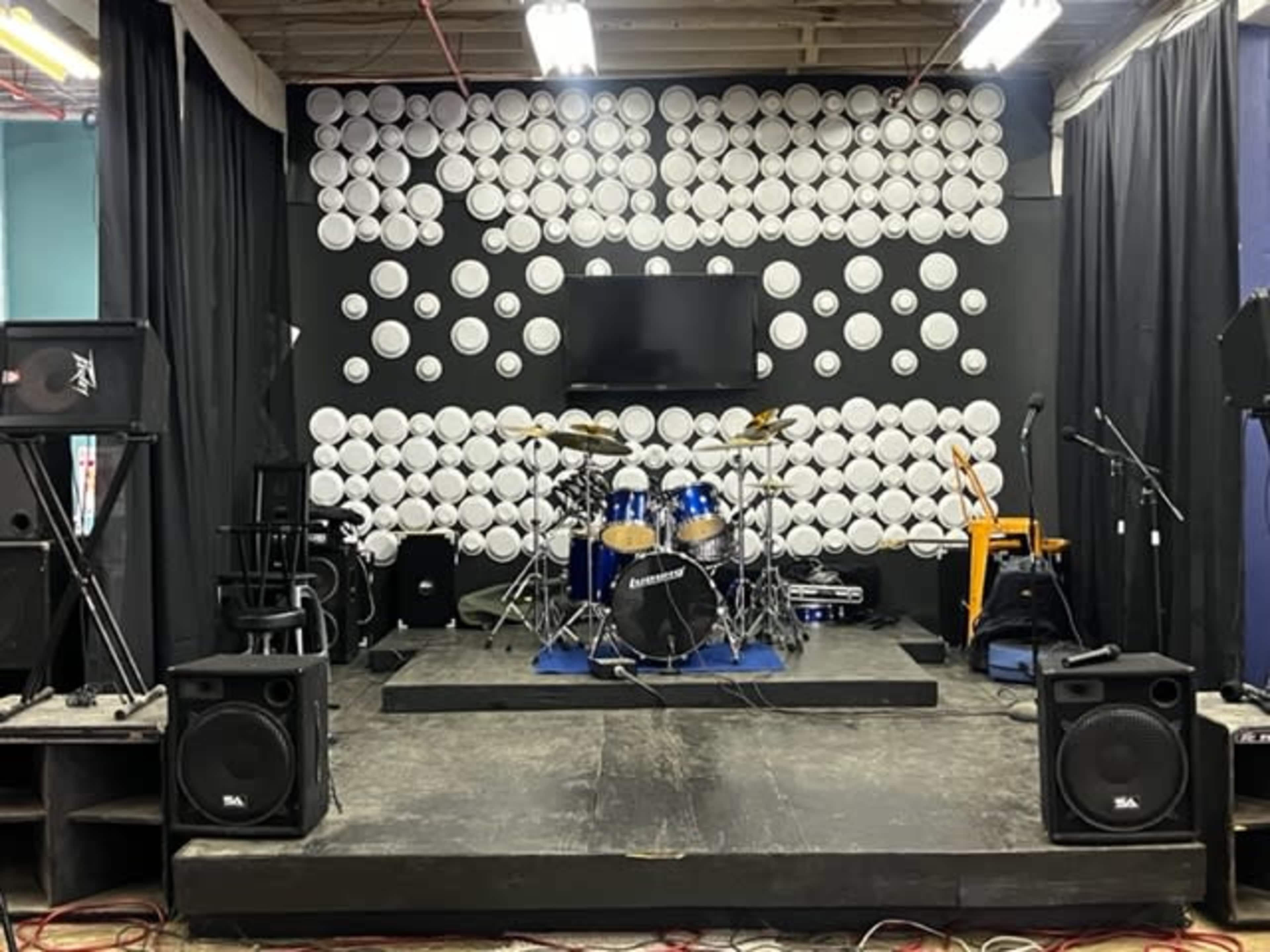 The image shows a stage setup with a drum kit, amplifiers, and instruments against a backdrop of white circular decorations.