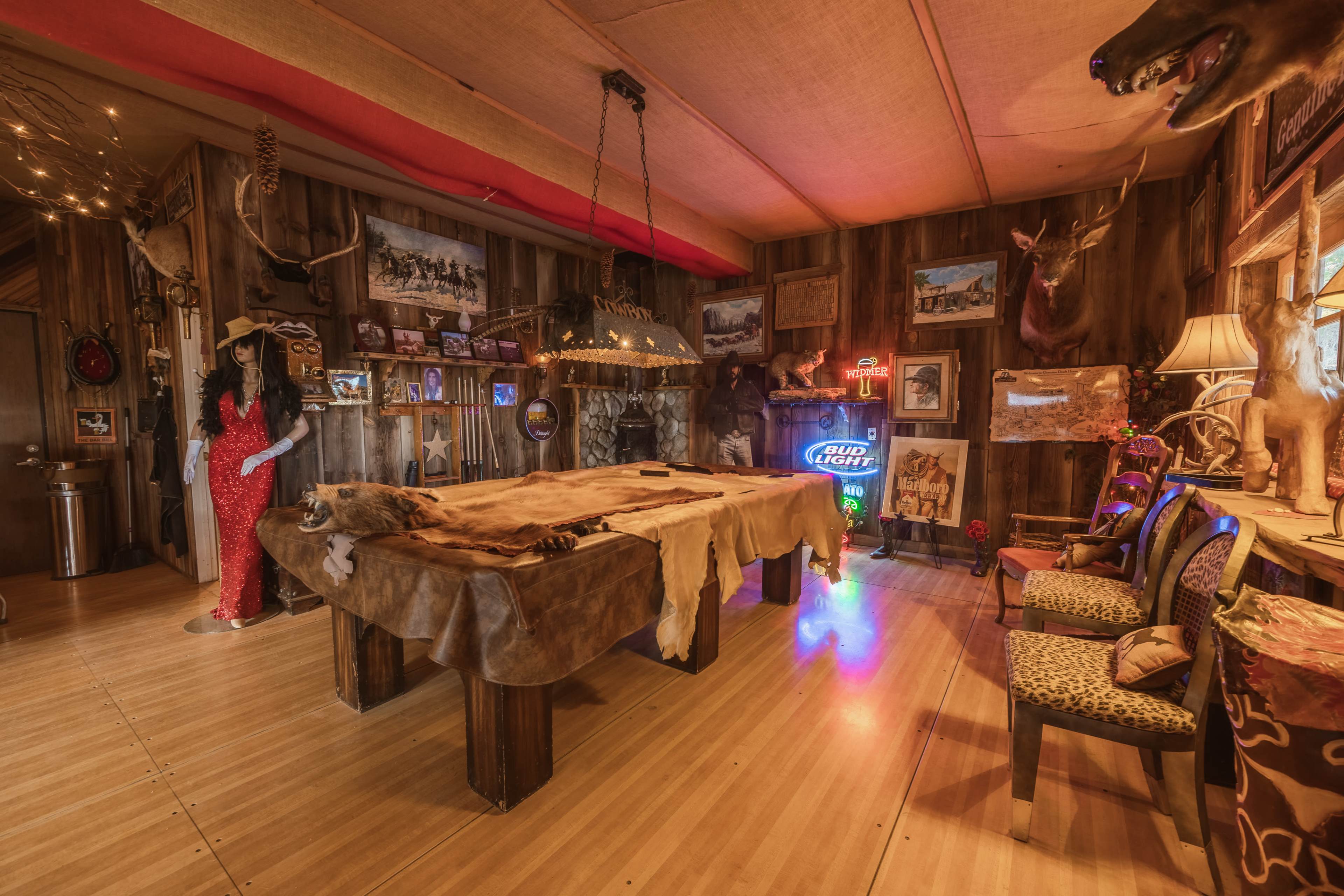 The image shows a rustic room with wooden paneling, featuring a large table covered with a fur hide, surrounded by chairs, and adorned with various mounted animal heads and framed photographs on the walls.