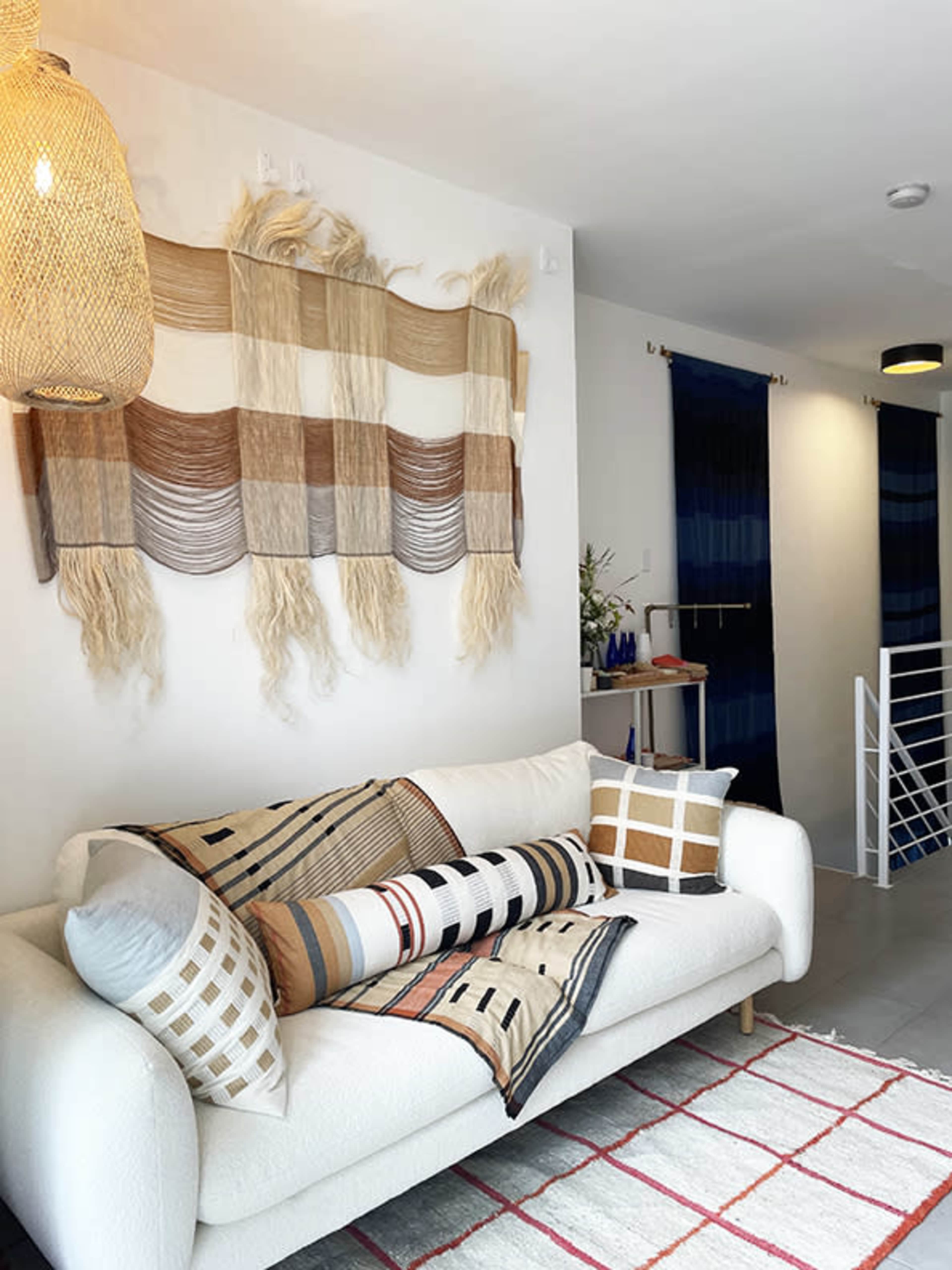 A modern living room features a white sofa adorned with patterned pillows, a textured wall hanging above, and colorful curtains in the background.