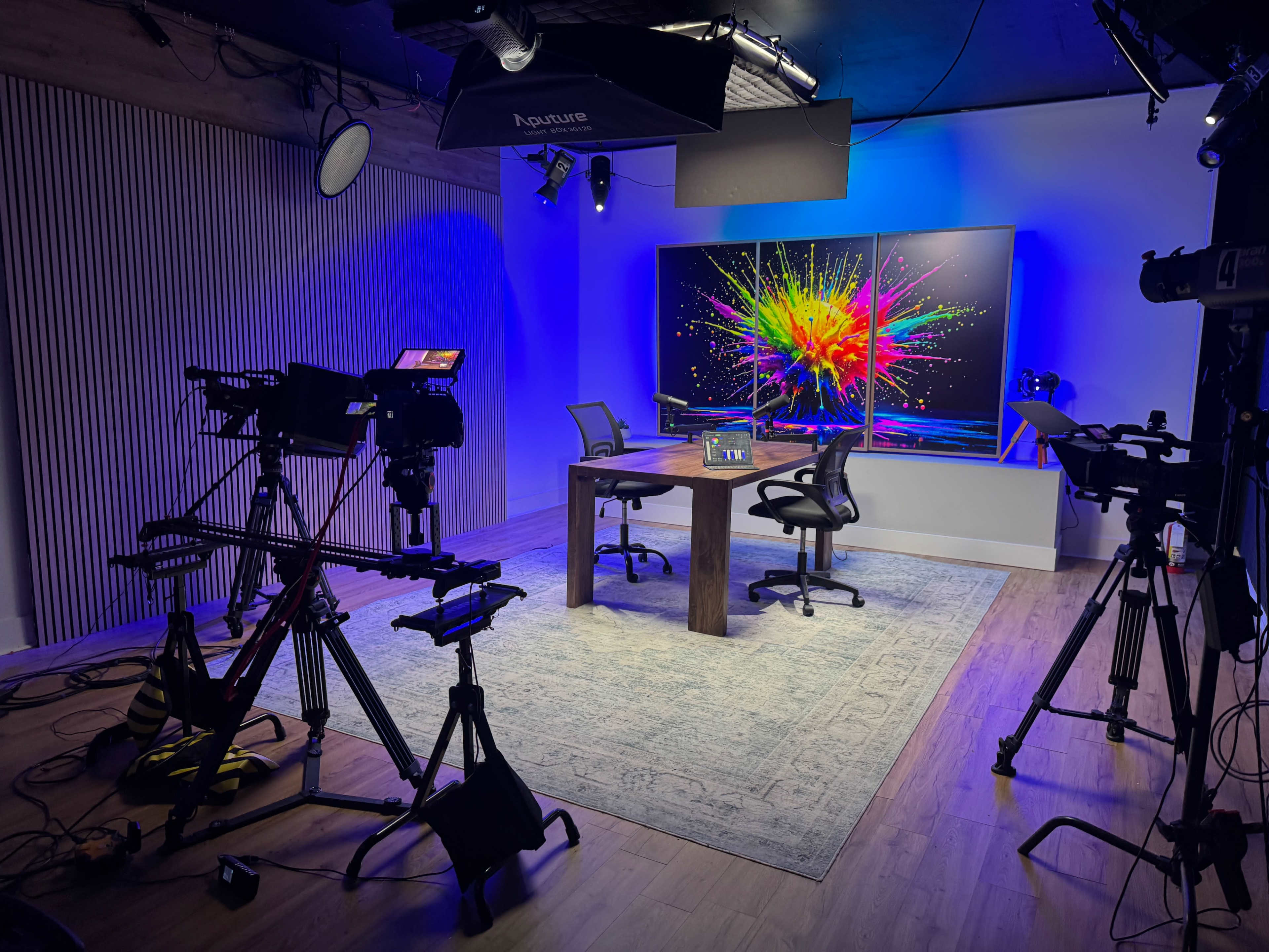 The image shows a modern studio setup with a central table and two chairs, surrounded by several cameras and lighting equipment, and a vibrant abstract artwork on the wall.
