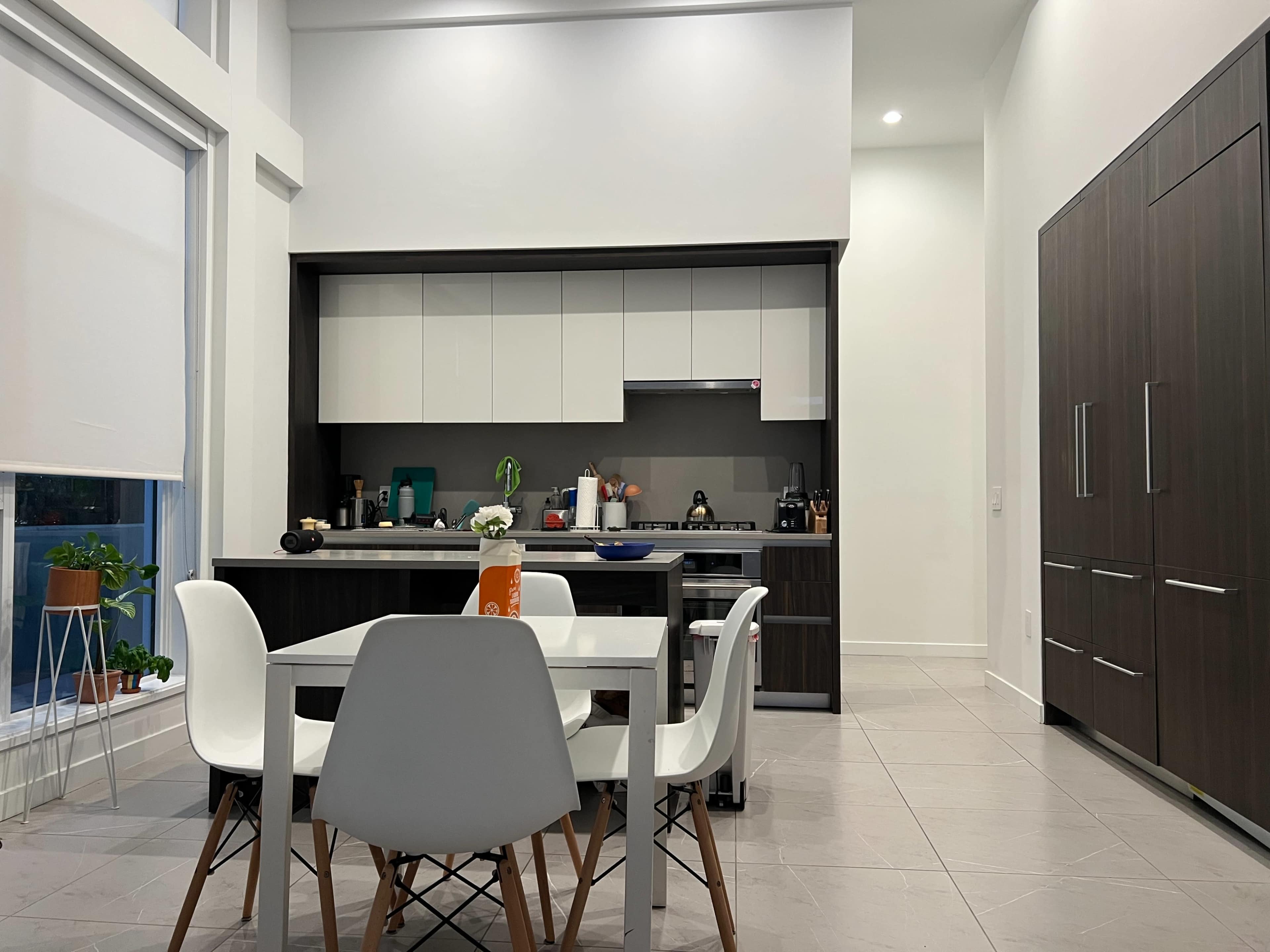 The image depicts a modern kitchen and dining area with a sleek white table surrounded by chairs, a dark wood cabinetry kitchen, and a bright, open space.