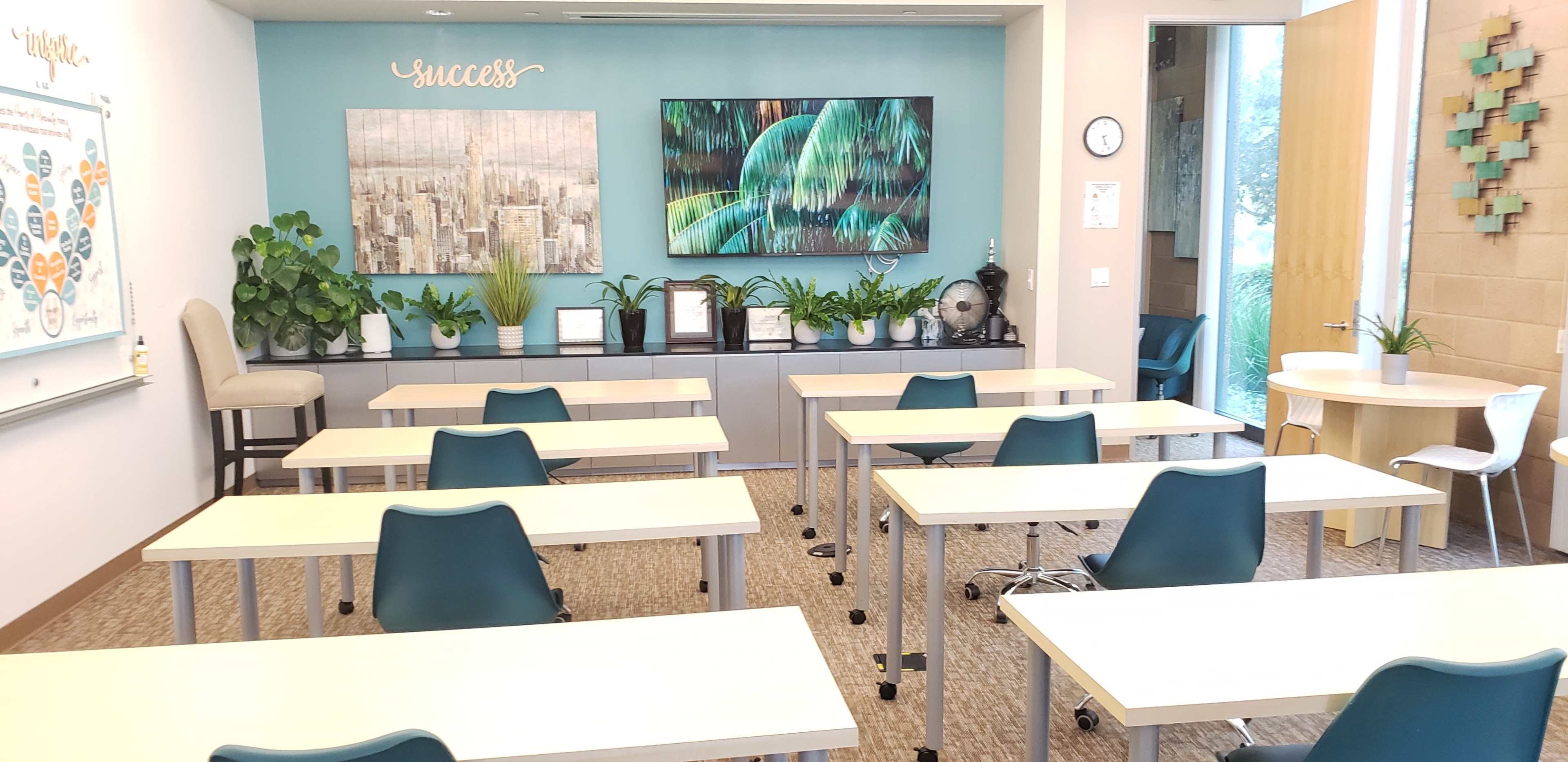 A modern classroom features several white tables and teal chairs arranged facing a wall adorned with two large artworks and potted plants, with a whiteboard on the left side.
