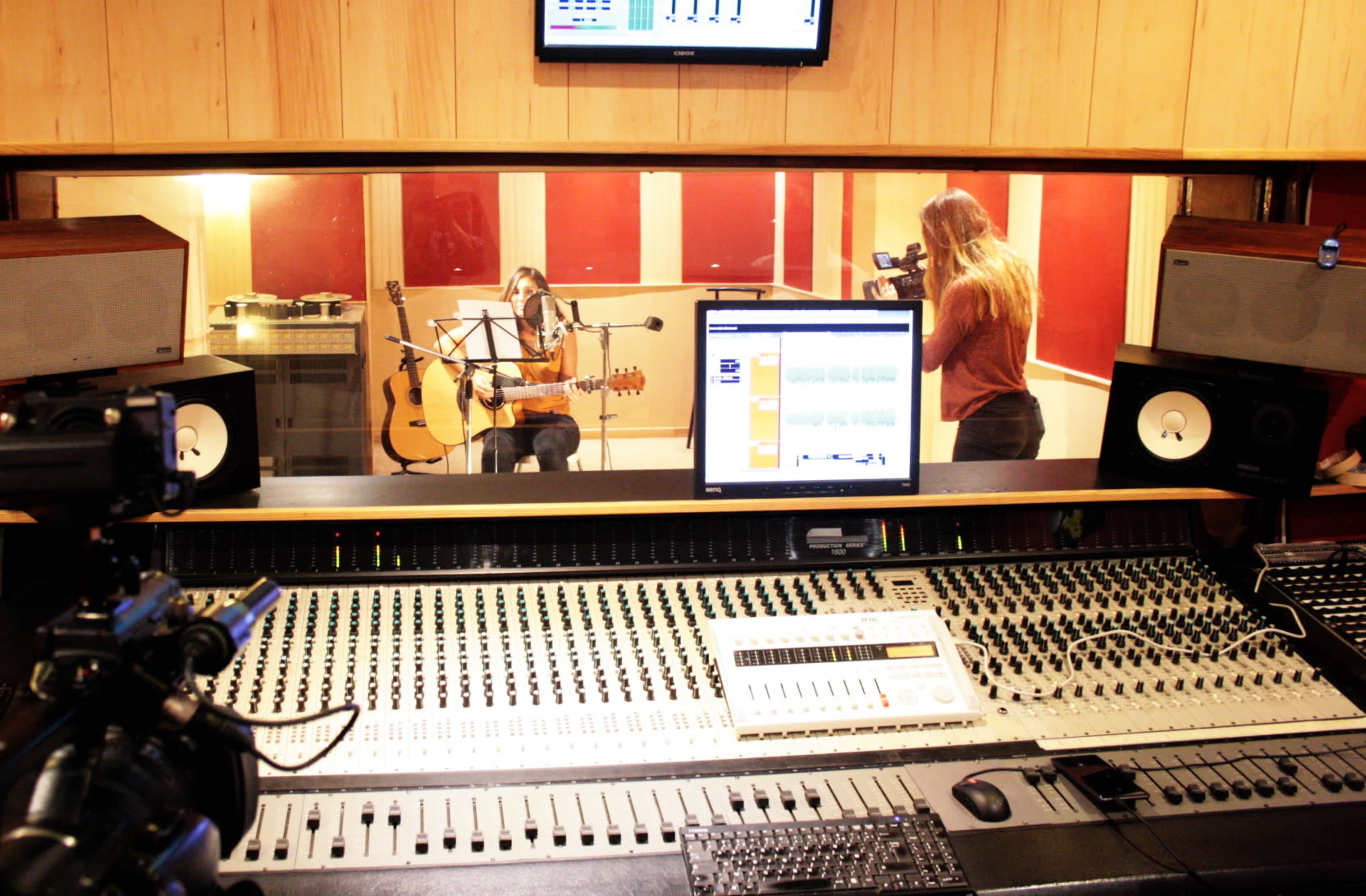 A musician plays an acoustic guitar in a recording studio while being filmed, with a sound mixing console in the foreground.
