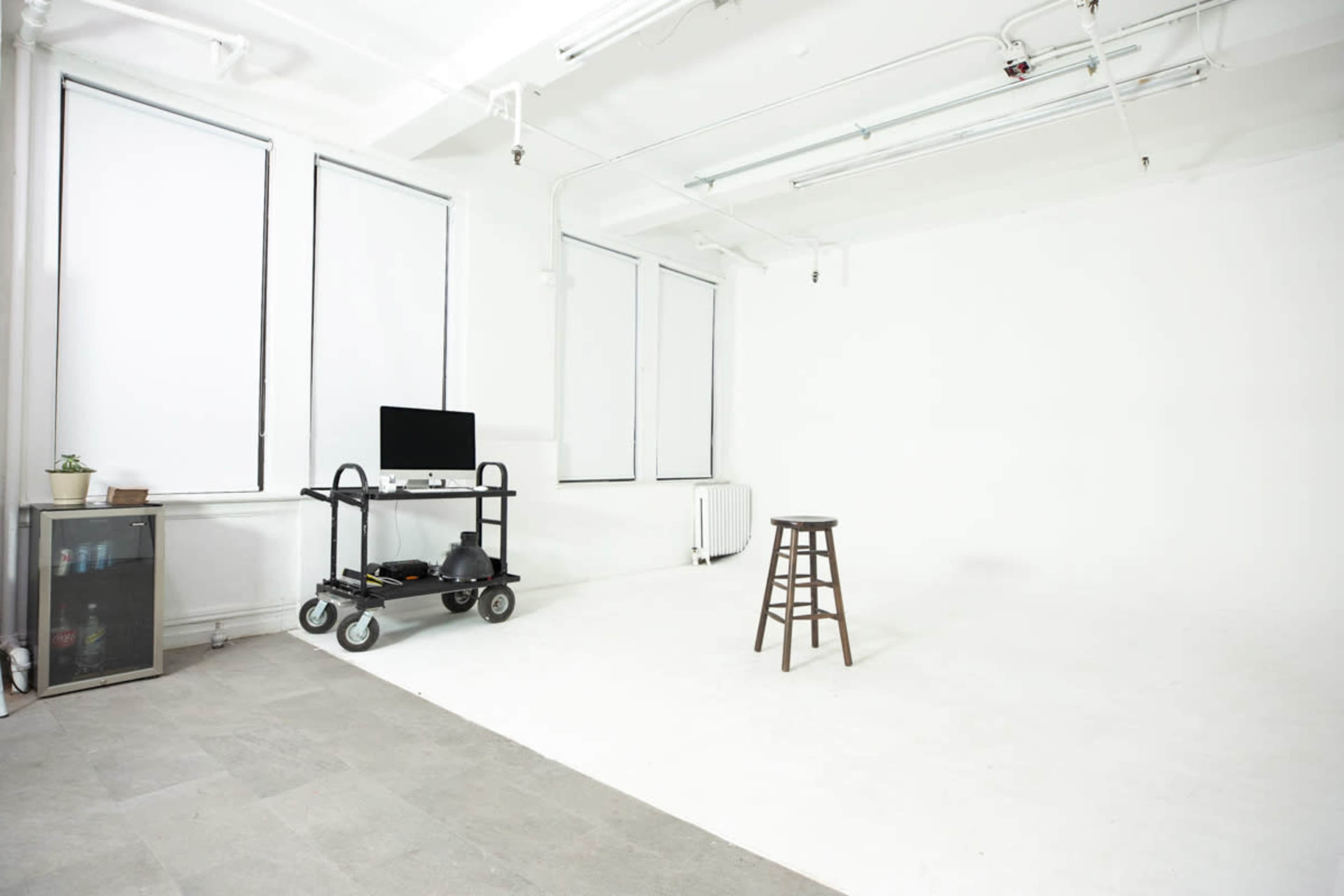 The image shows a minimalistic studio with white walls, a wooden stool, a cart with a monitor, and large windows.