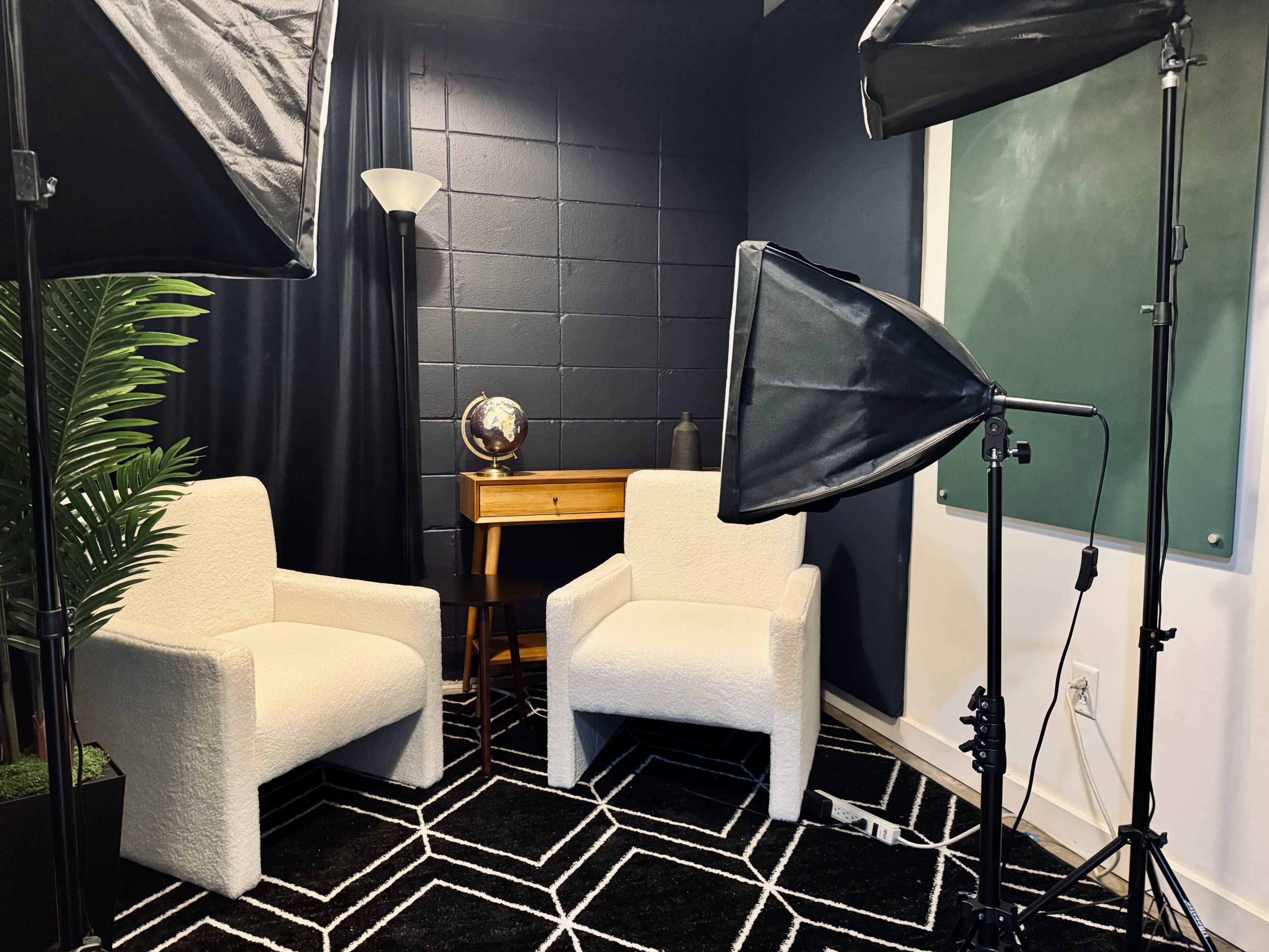 The image shows a small studio setup featuring two white chairs, a wooden table with a globe, softbox lights, and a dark wall with a chalkboard.