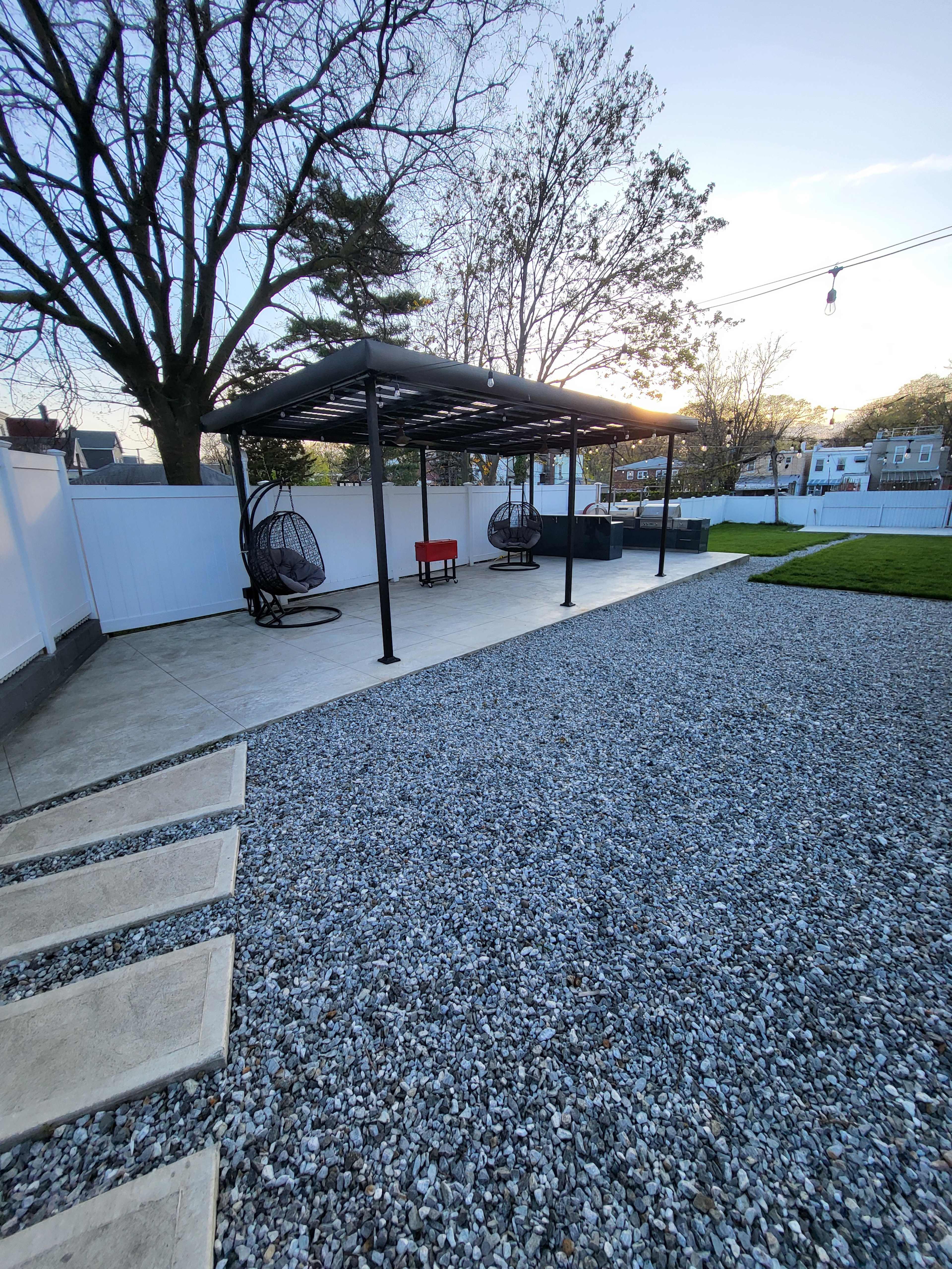 A backyard patio with a gravel area, a few hanging chairs, and a covered section with a red table.