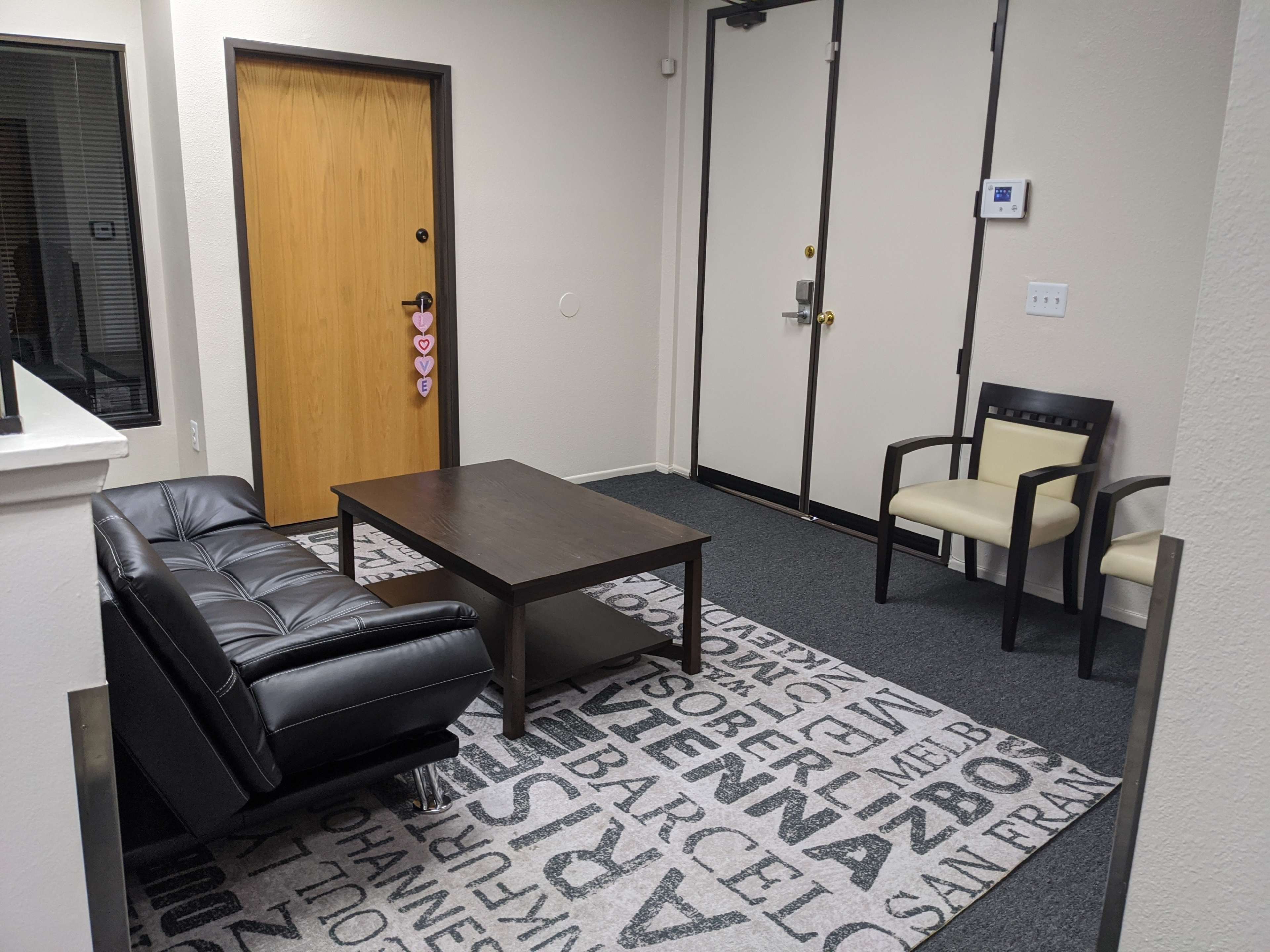 A small waiting area with a black sofa, a coffee table, and two chairs on a patterned carpet near a door.