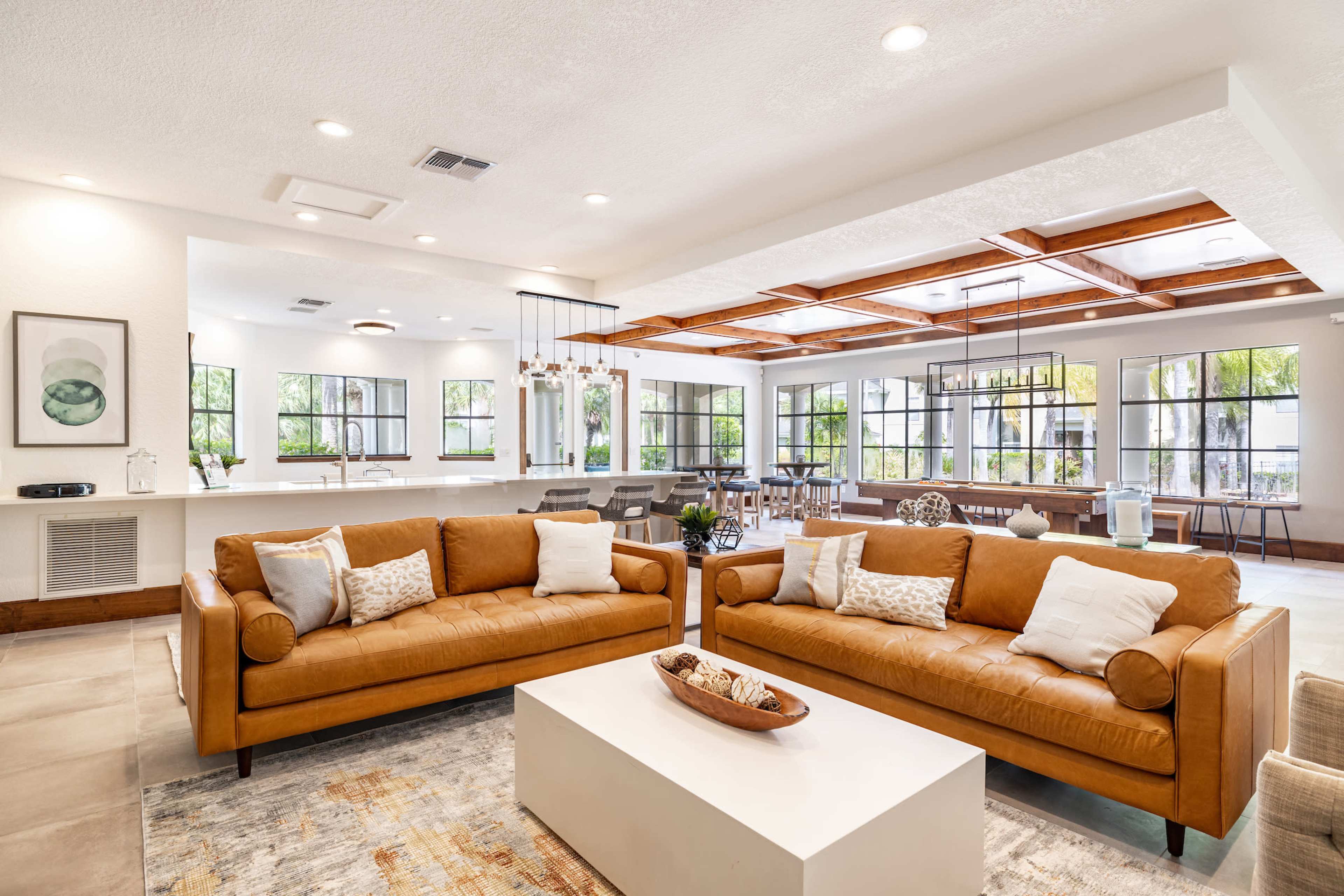 A modern living room furnished with two brown leather sofas facing a white coffee table, surrounded by bright windows and an open dining area in the background.