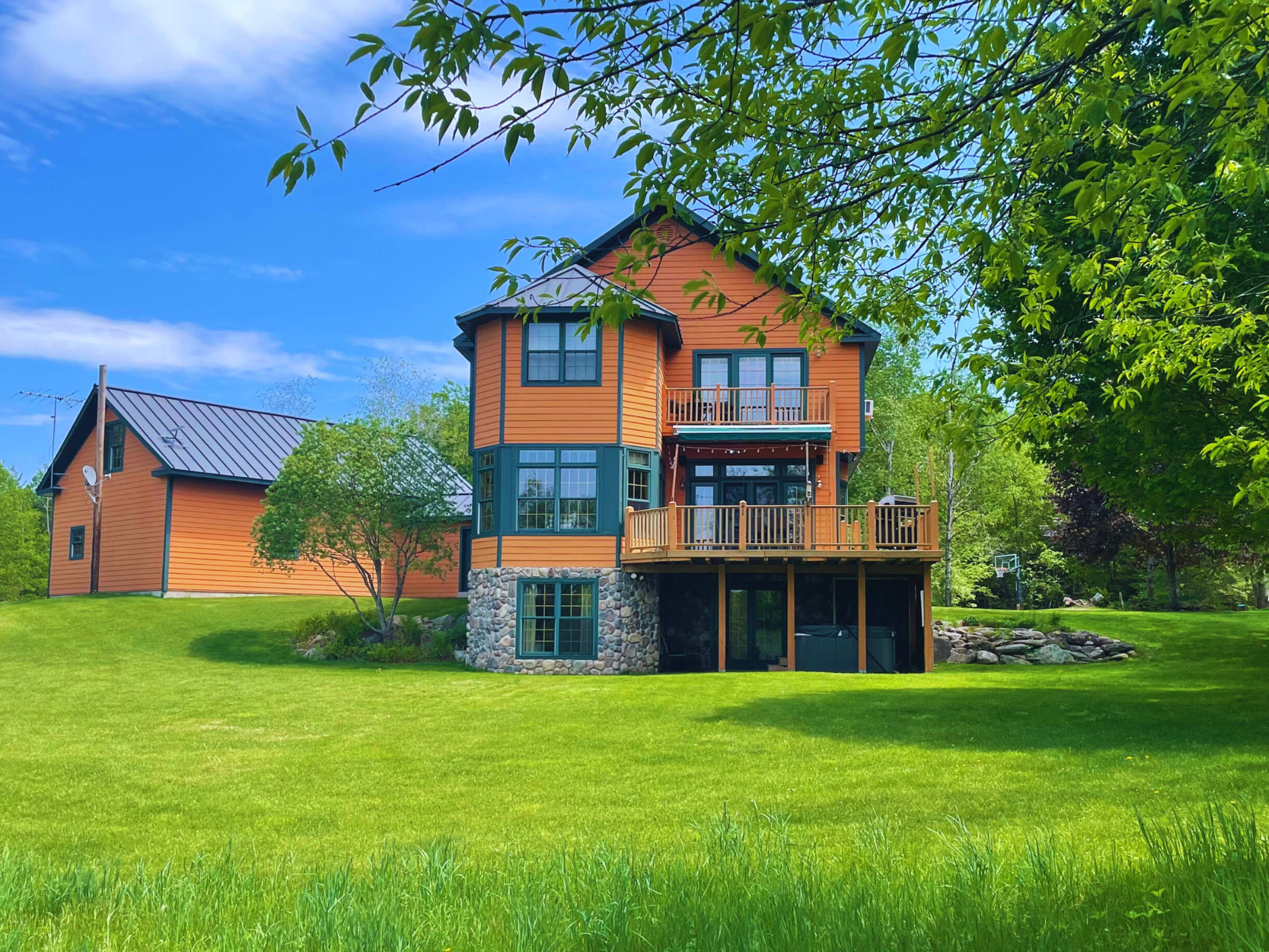 A large, orange house with a stone foundation and multiple decks is set on a grassy lawn surrounded by trees.