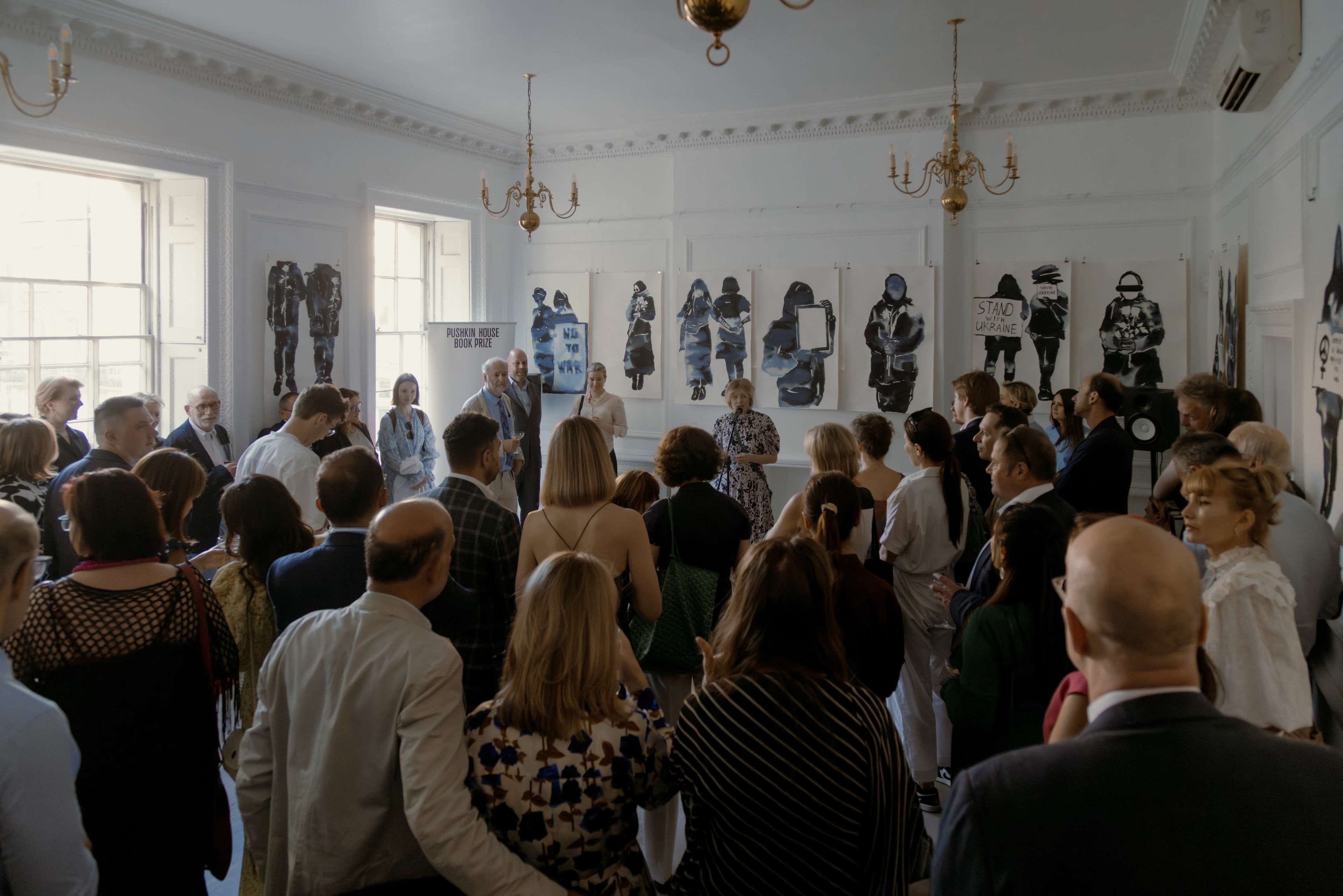 A large group of people gathers in a well-lit room adorned with black-and-white artwork on the walls, while an individual stands in front to address the audience.