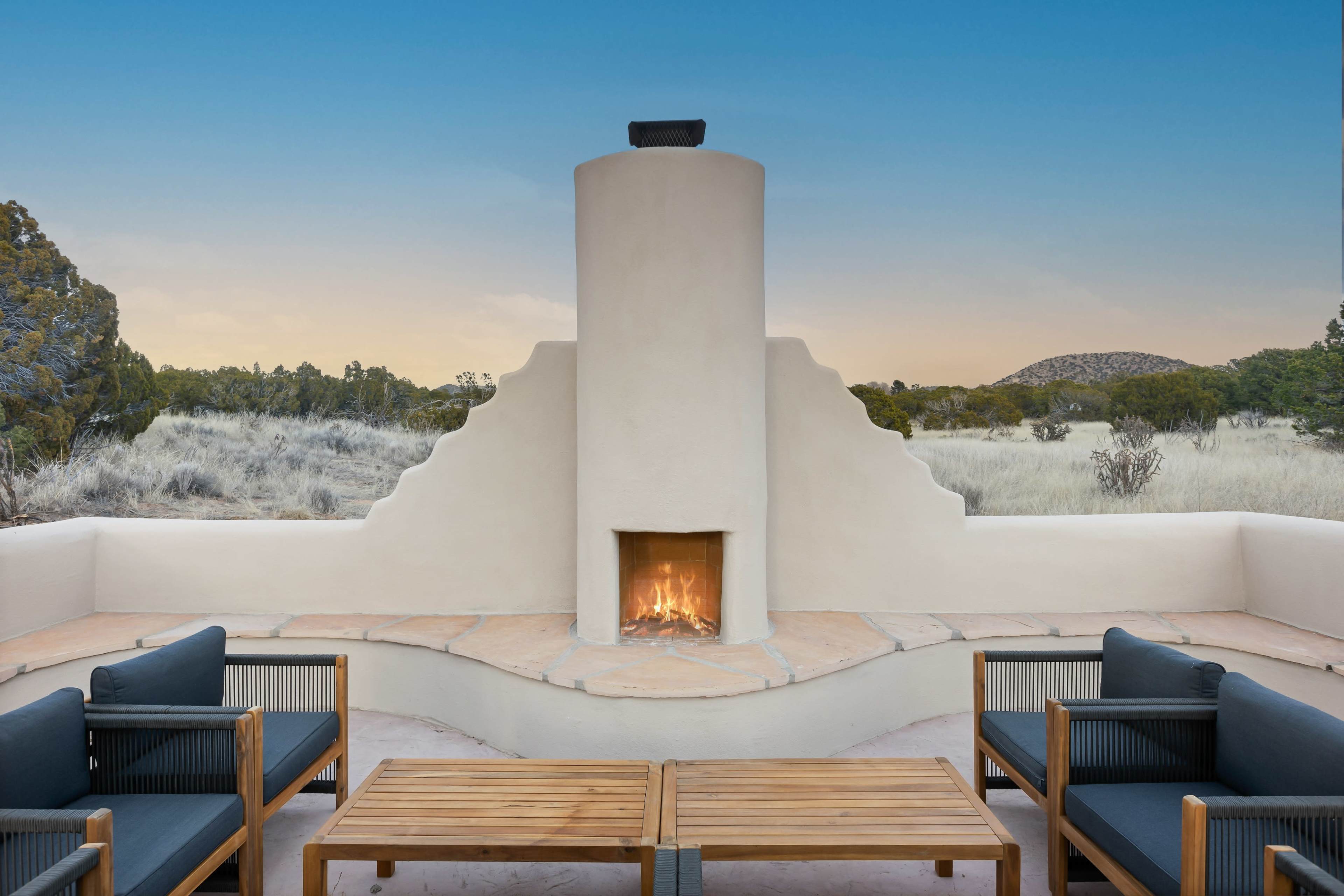 A modern outdoor seating area features a central fireplace surrounded by wooden furniture, with a backdrop of desert vegetation and a clear sky.