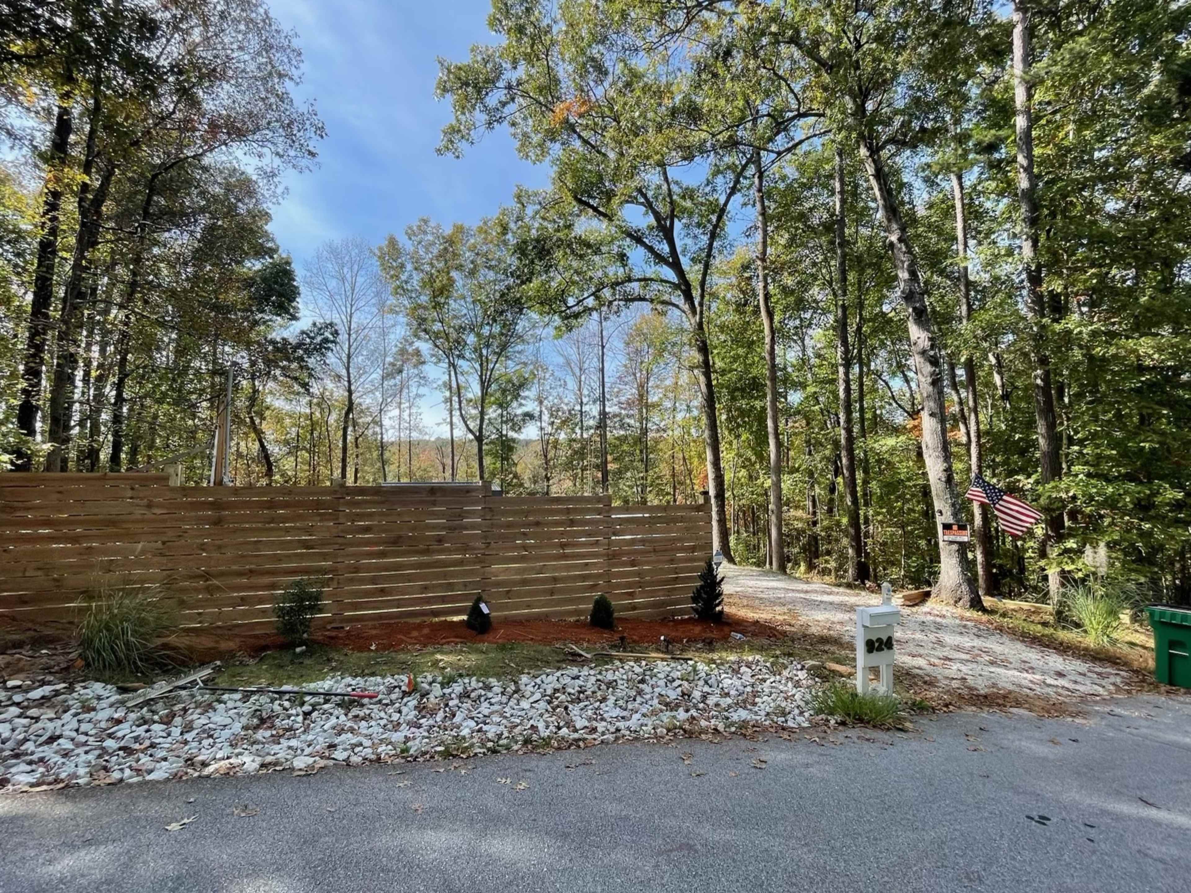 A gravel driveway leads to a wooden fence surrounding a property, with neatly arranged plants and a small American flag visible nearby.