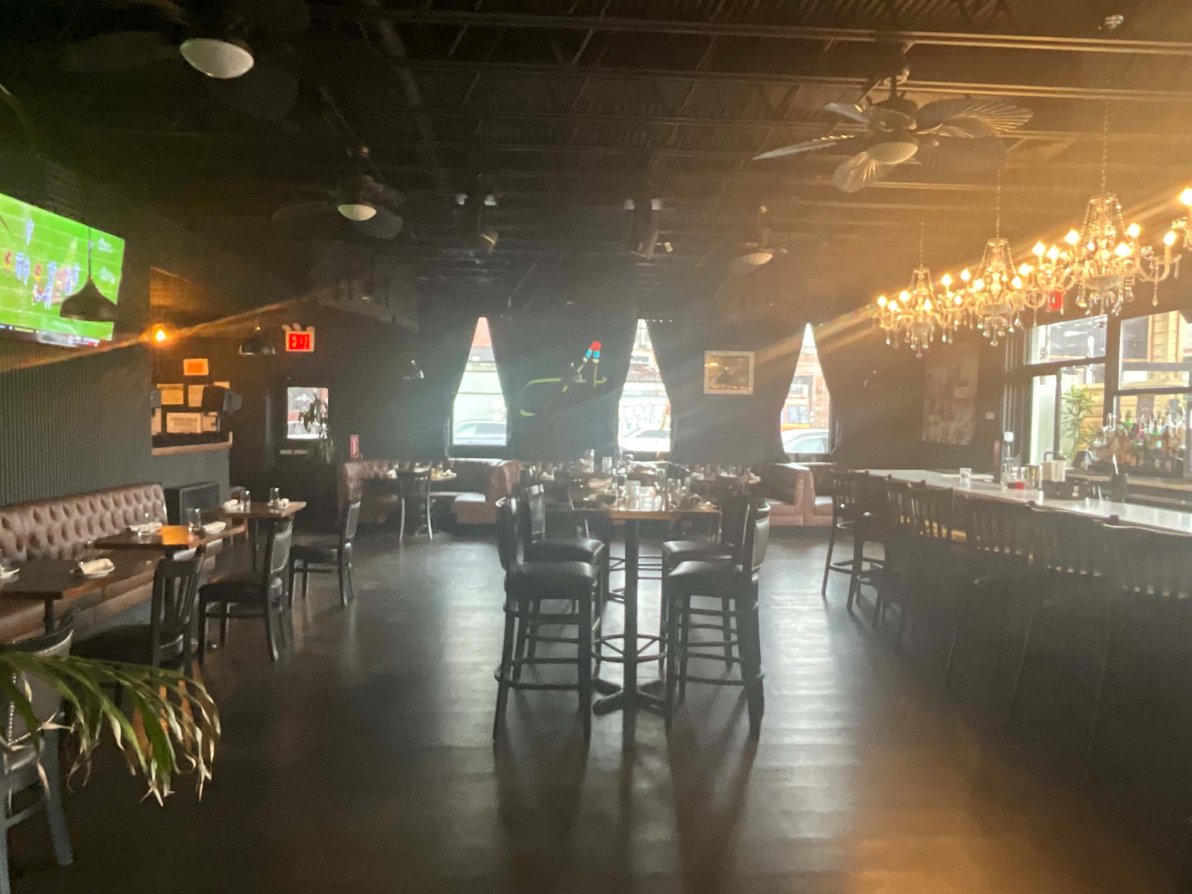 The image shows a dimly lit restaurant interior with tables set for dining and a bar area, featuring a chandelier and a large television screen in the background.