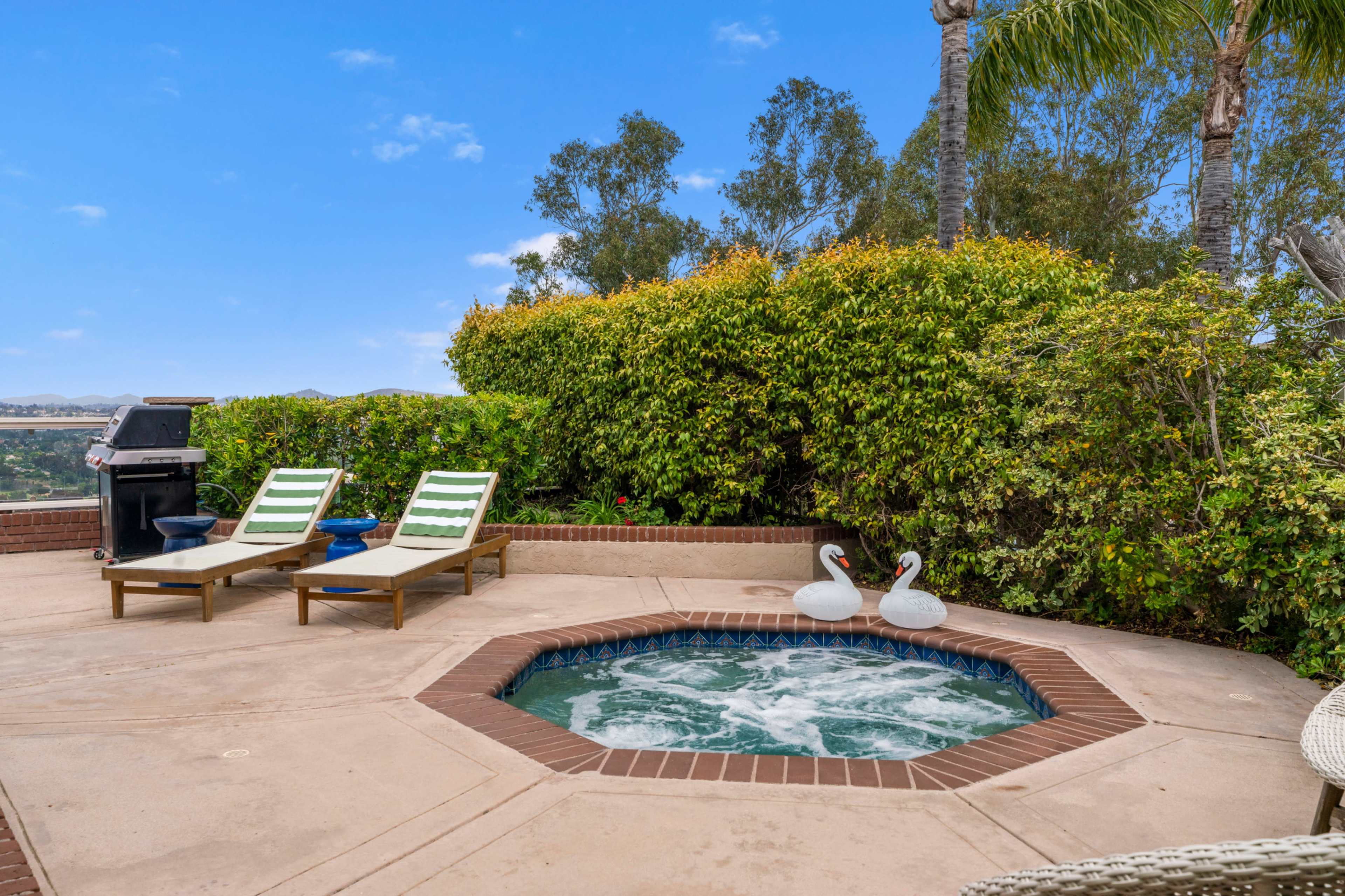 The image shows a patio area with two lounge chairs, a hot tub, and decorative swan figures surrounded by green shrubs.
