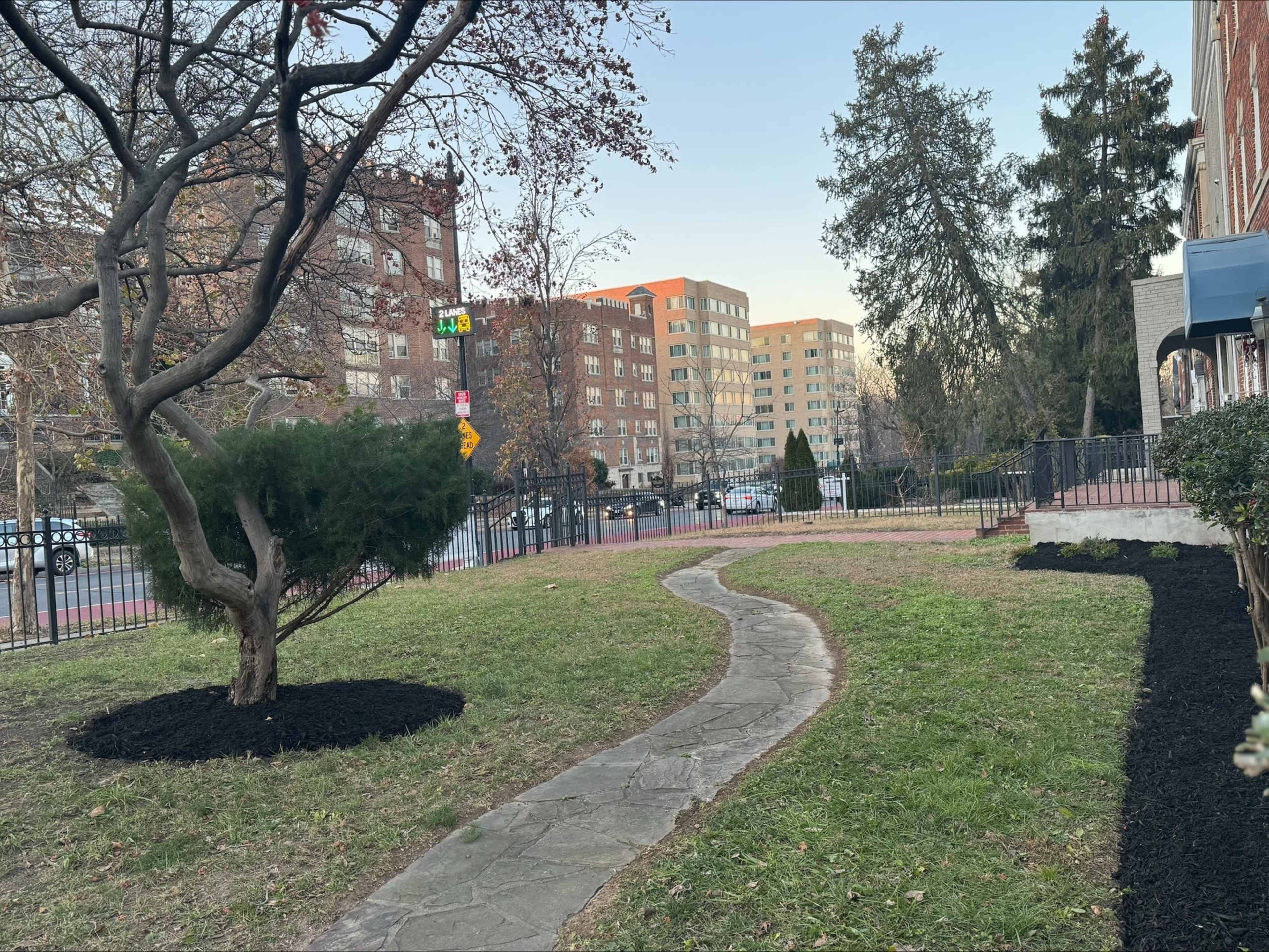 The image shows a landscaped area with a winding stone path, a tree, and freshly laid mulch, bordered by a black fence and residential buildings in the background.