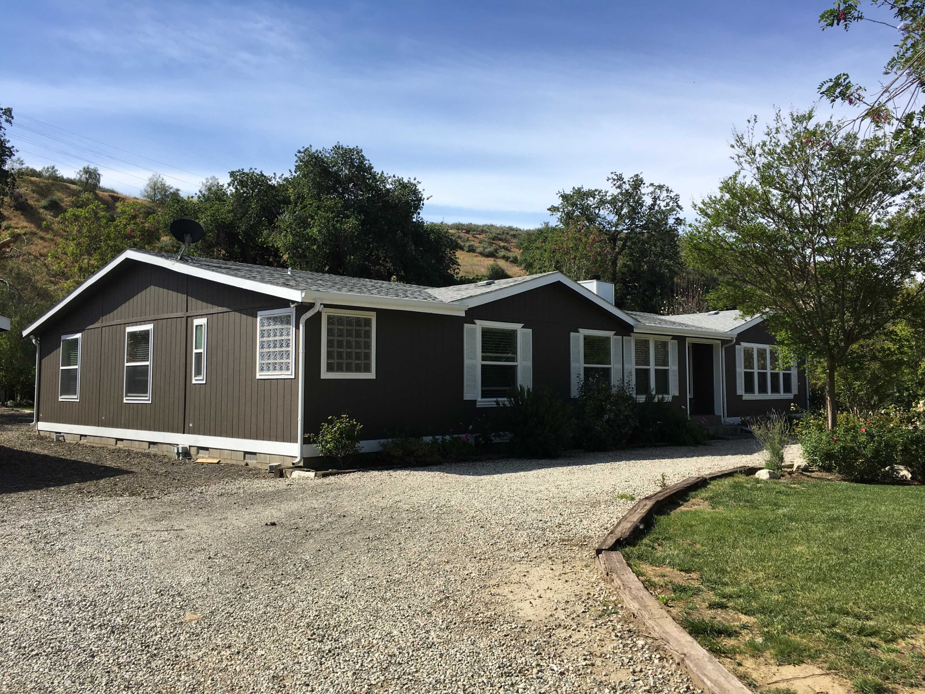 A single-story house with a dark gray exterior and a gravel driveway, surrounded by trees and landscaping.