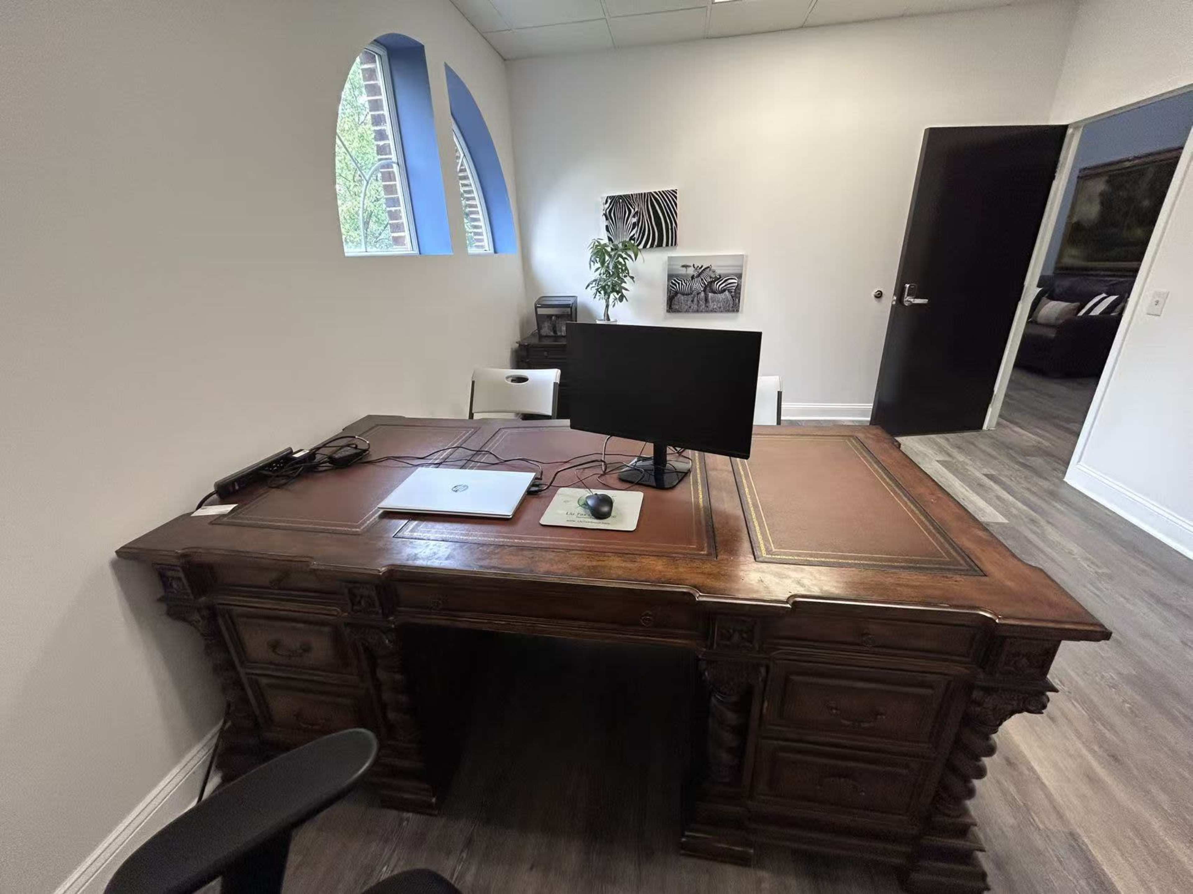 The image shows a wooden desk with a computer monitor and a laptop, situated in a well-lit office with a partially opened door leading to another room.