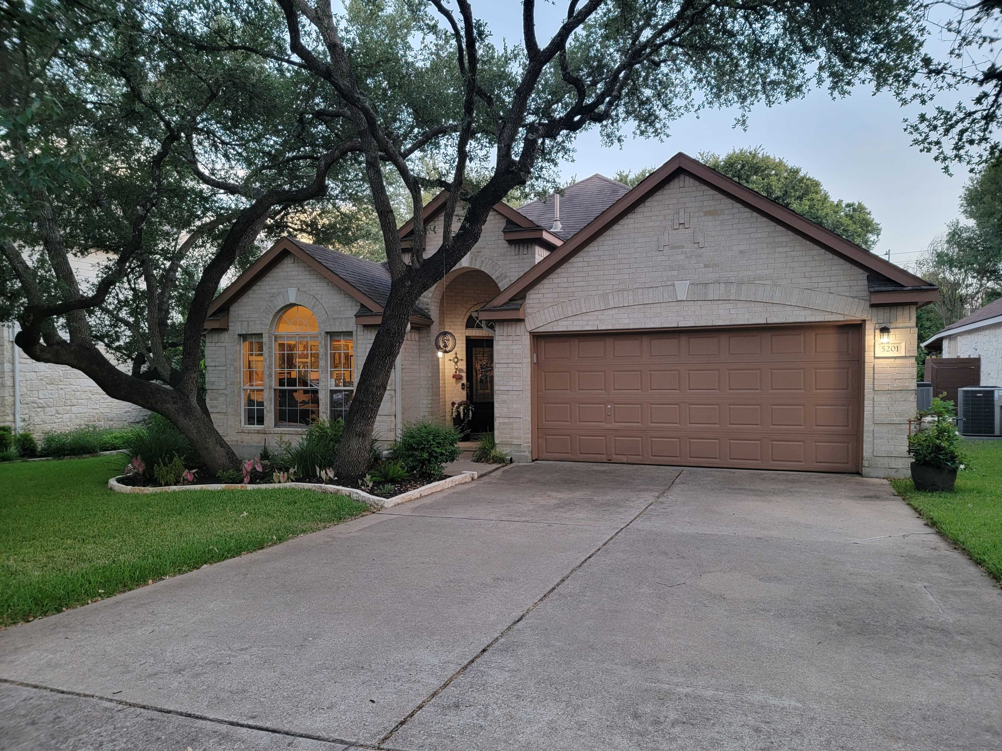A single-story brick house with a garage sits on a neatly landscaped lot, featuring large trees and a well-maintained lawn.