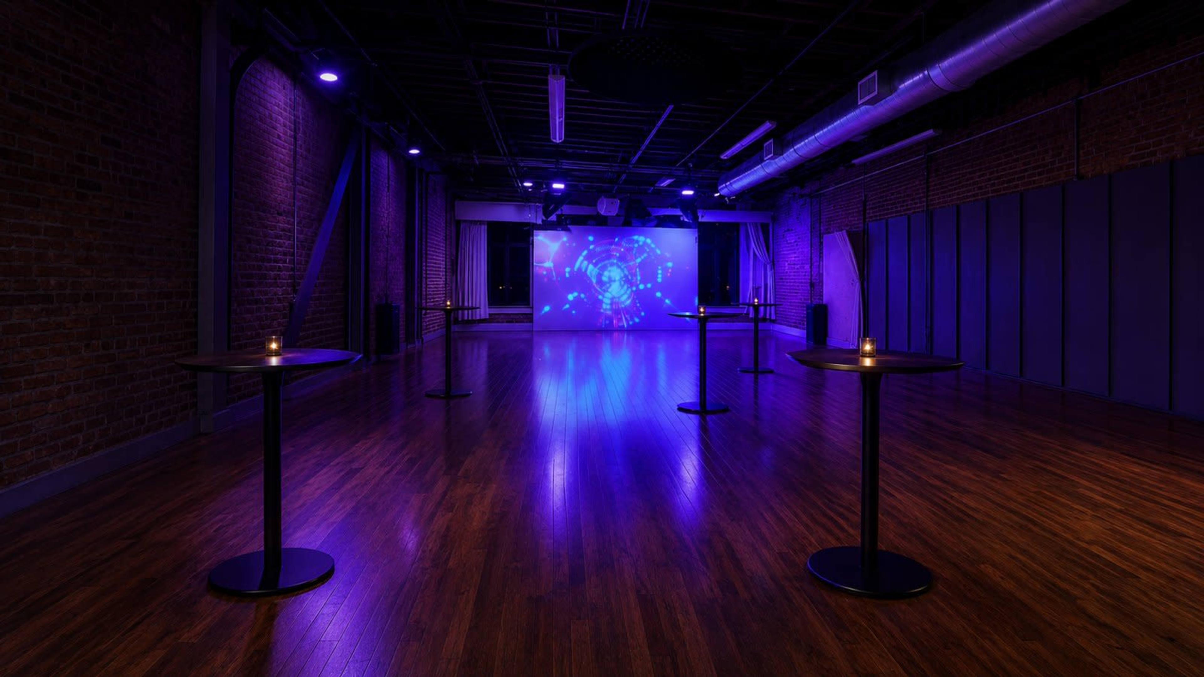 The image shows a spacious, dimly lit event venue with wooden floors and purple lighting, featuring several small round tables set up in a rectangular arrangement.