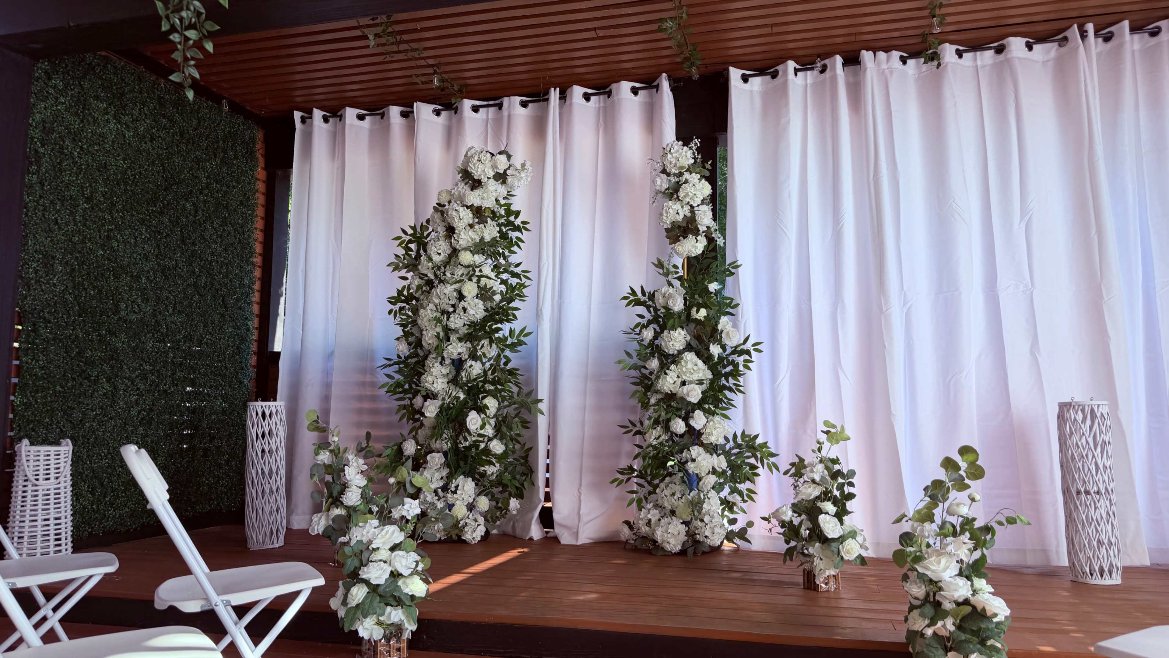 The scene features a decorated stage with tall floral arrangements and white drapery, flanked by smaller flower displays on the floor.
