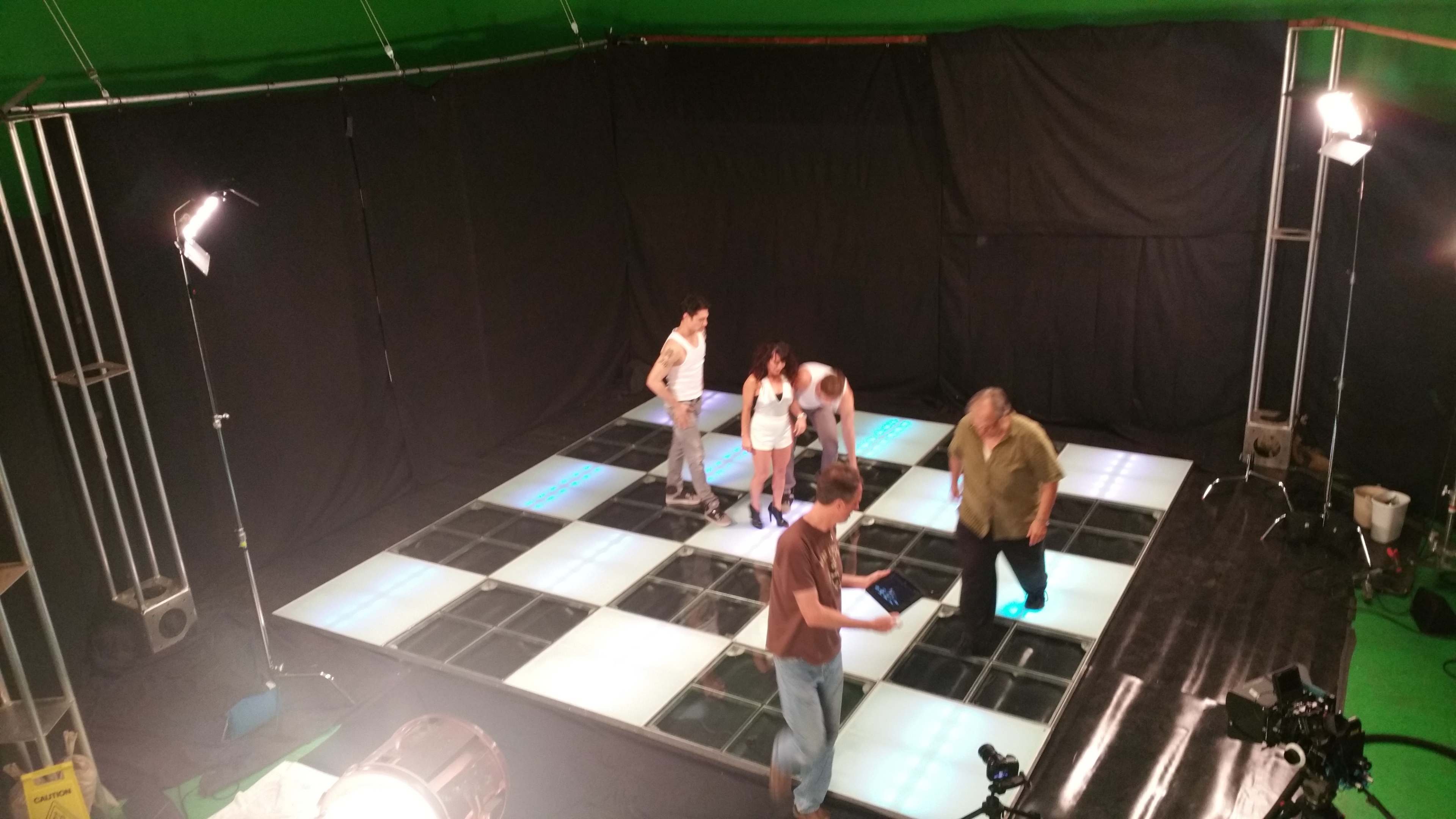 A group of five people stands on a large illuminated grid floor in a studio with a green screen backdrop.