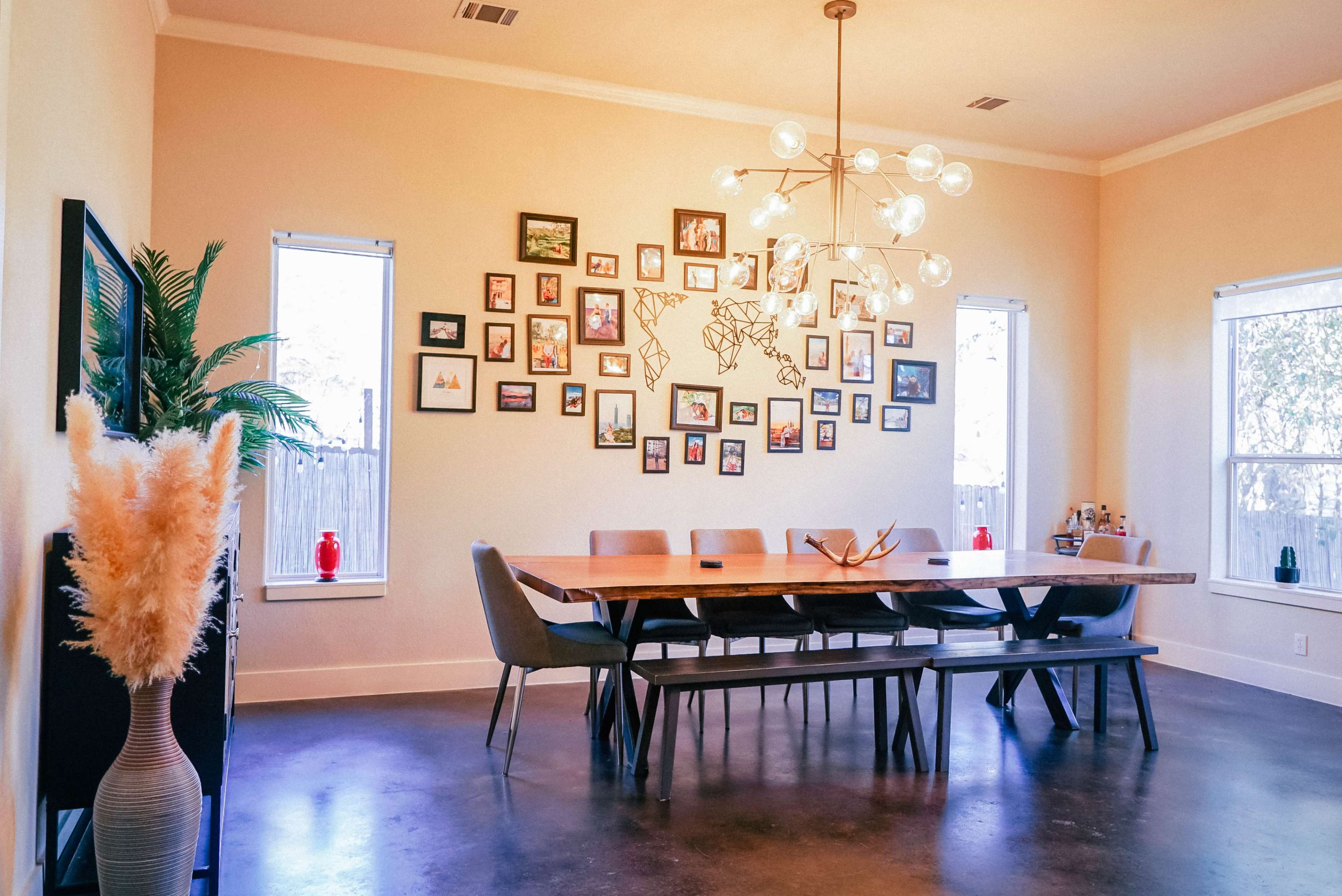 A spacious dining room features a long wooden table surrounded by gray chairs, with a gallery of framed photos on the wall and large windows letting in natural light.