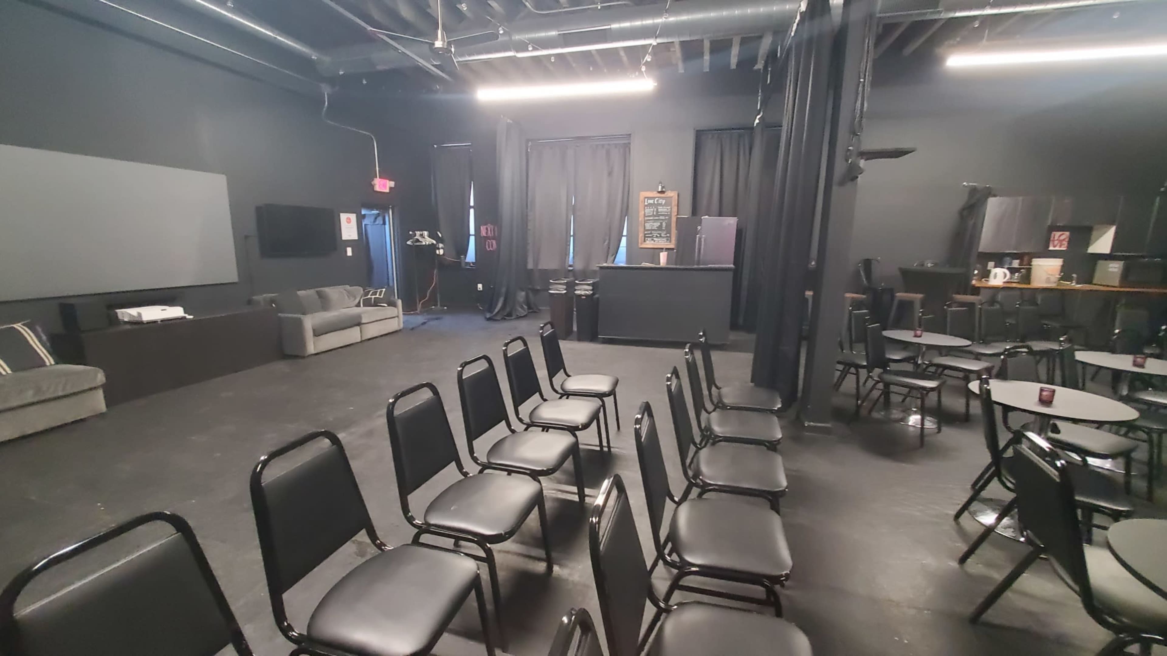The image shows a black-walled performance space with rows of black chairs facing a bar area, a sofa in the corner, and dim lighting.